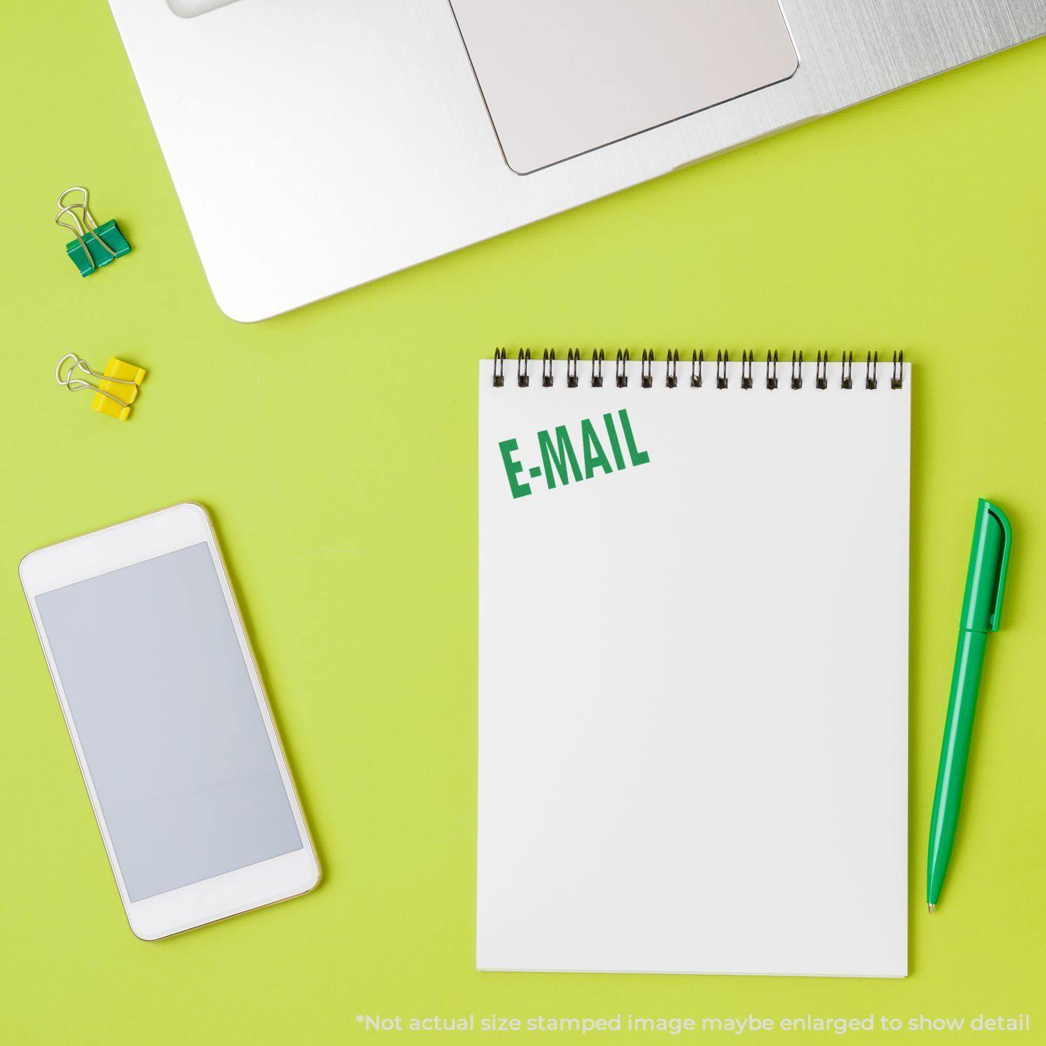 Self Inking E Mail Stamp used on a white notepad next to a green pen, smartphone, laptop, and yellow and green binder clips.