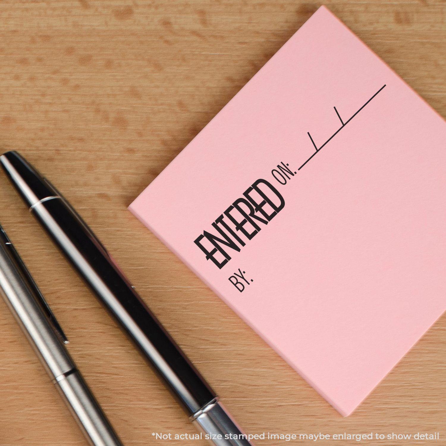 Slim Pre-Inked Entered On Stamp on a pink sticky note next to two pens on a wooden surface.