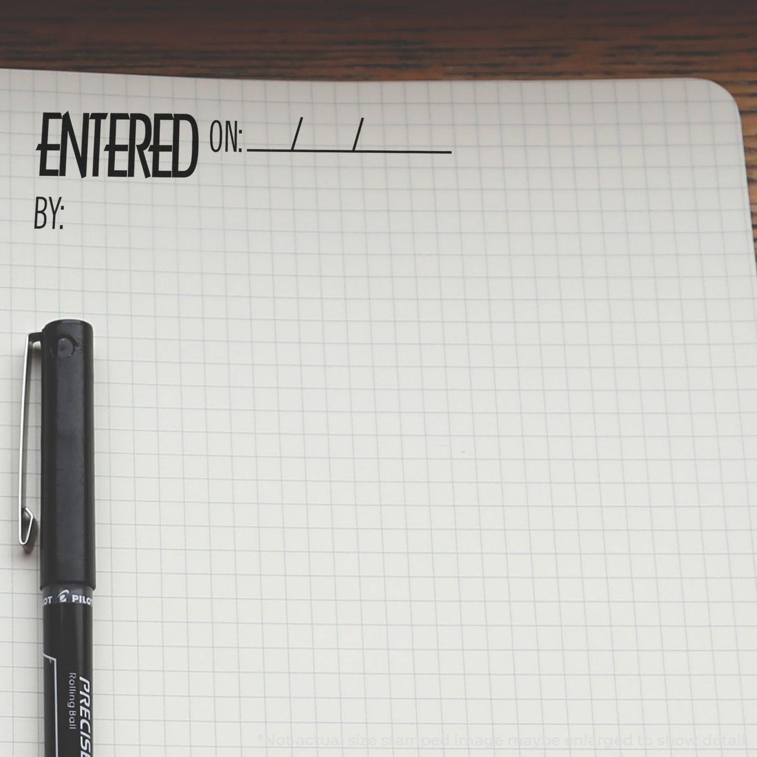 A Large Self Inking Entered On Stamp is used on a grid notebook page with a black pen placed beside it.