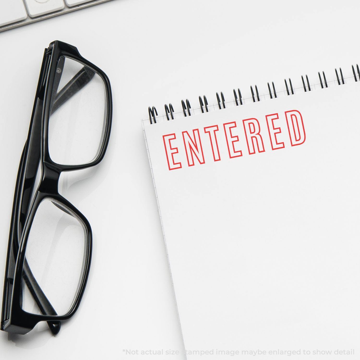 Large Pre-Inked Entered Outline Stamp in red ink on a white notepad, next to black glasses and a keyboard.