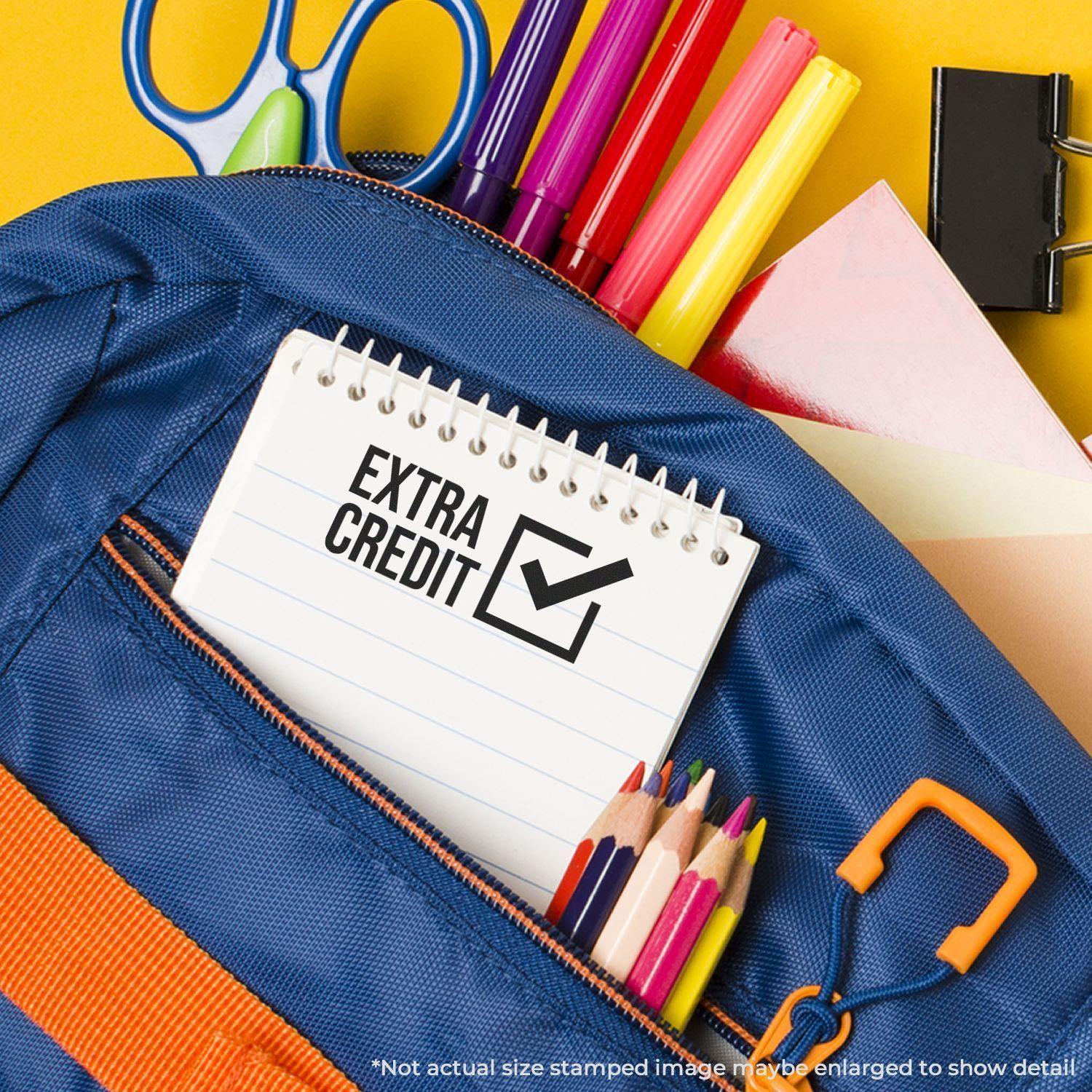 Slim Pre-Inked Extra Credit Stamp on a notepad inside a blue backpack with colored pencils, markers, scissors, and paper clips.