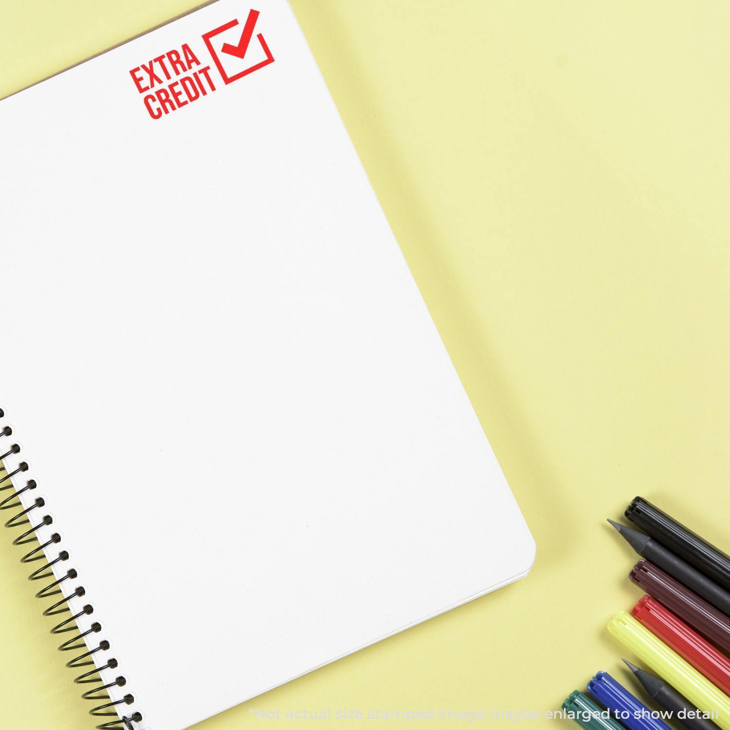 A notebook stamped with Extra Credit in red ink using the Extra Credit Rubber Stamp, with pens and markers on a yellow background.