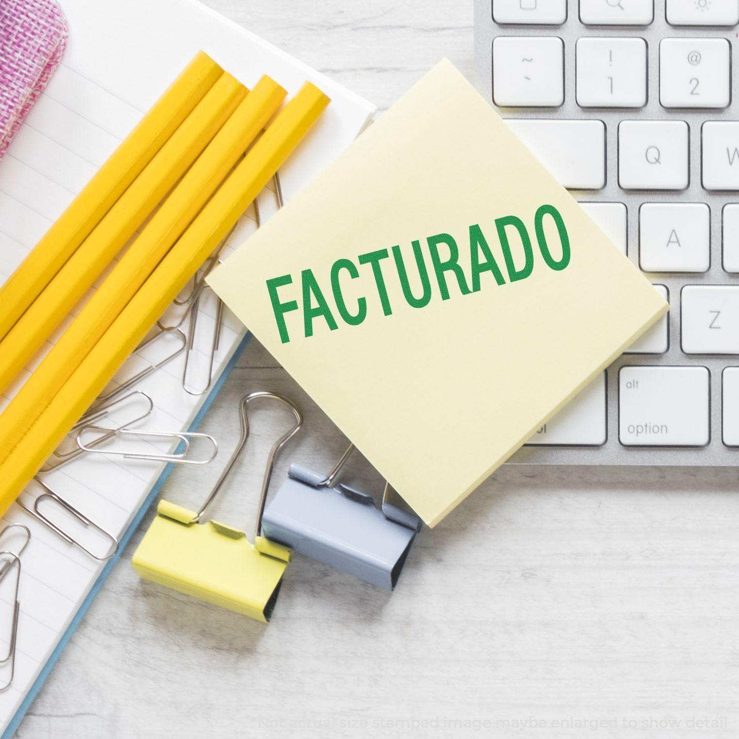 Large Facturado Rubber Stamp on a yellow sticky note, surrounded by pencils, paper clips, binder clips, and a keyboard on a desk.