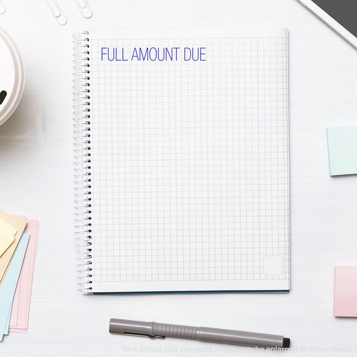 Large Self Inking Full Amount Due Stamp used on a spiral notebook with grid paper, surrounded by office supplies on a white desk.