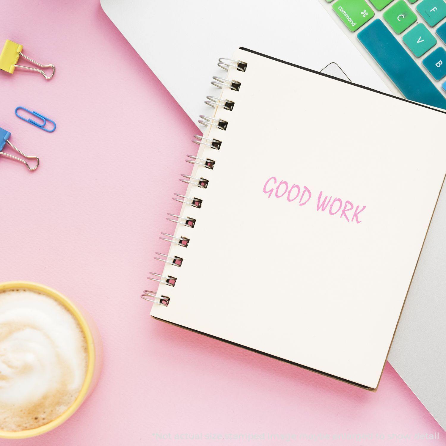 Good Work Rubber Stamp used on an open notebook, placed on a pink desk with a laptop, coffee, and colorful paper clips nearby.