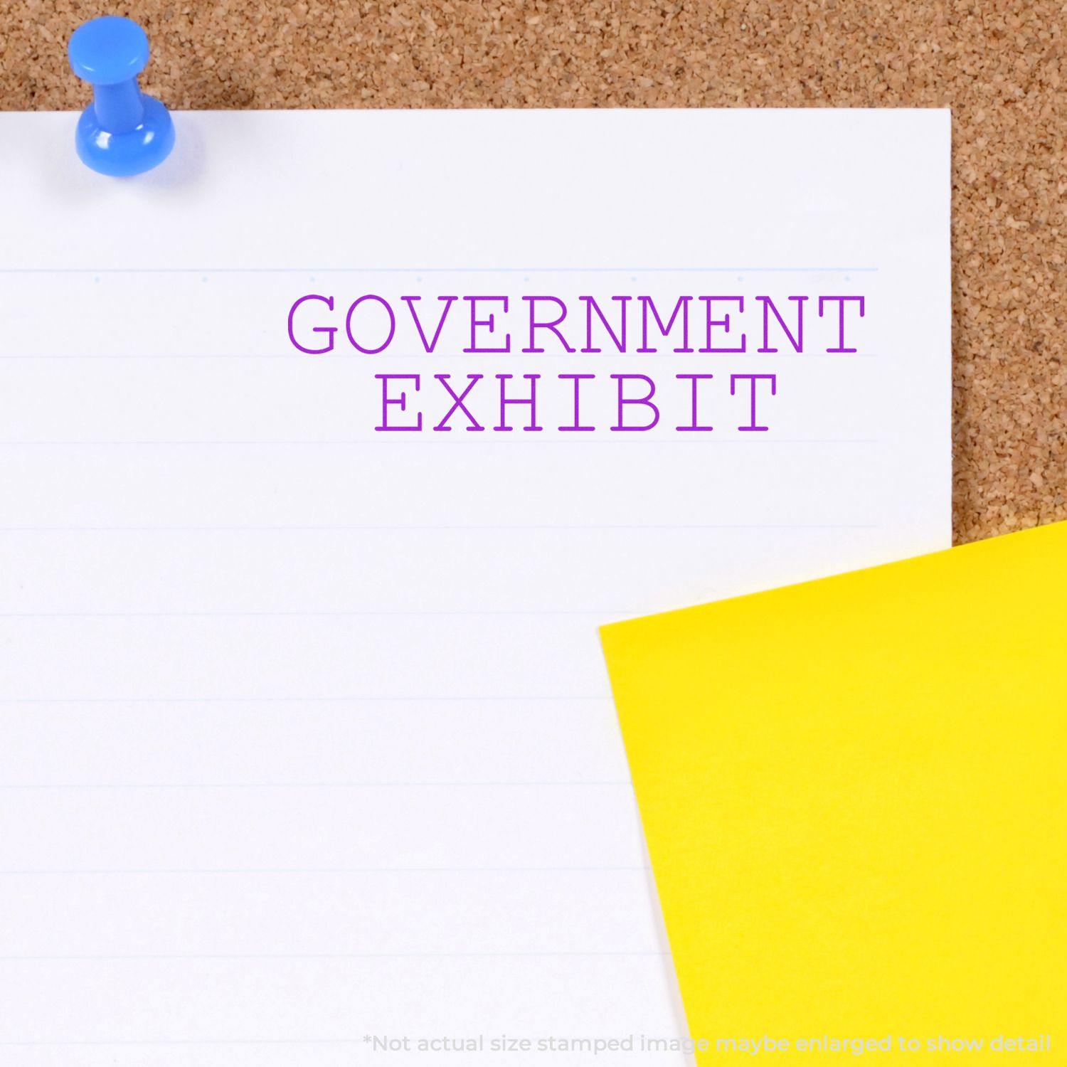 Self Inking Government Exhibit Stamp in purple ink on a white paper pinned to a corkboard, with a yellow paper partially visible.