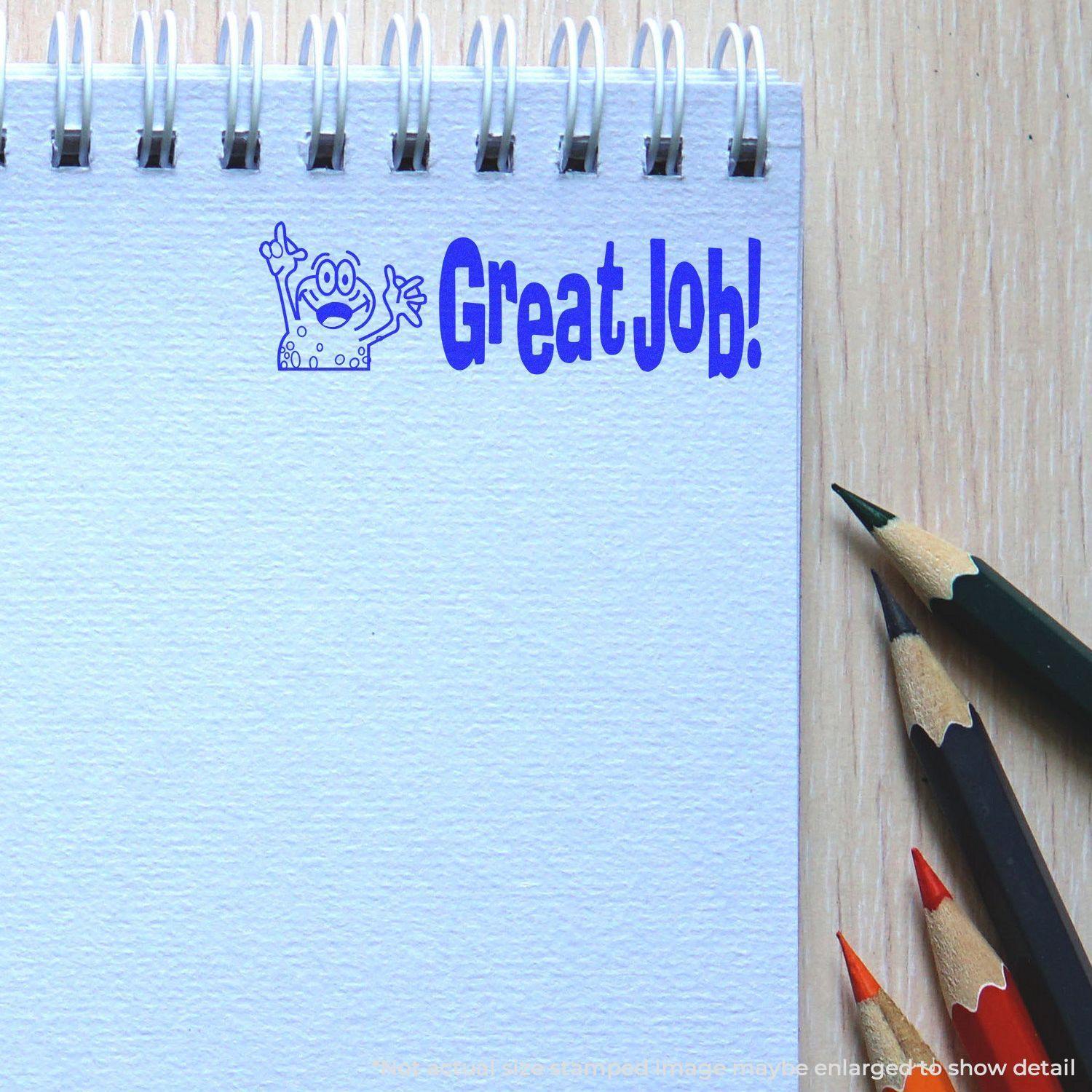 Large Self Inking Great Job Stamp in blue ink on a spiral notebook, with colored pencils nearby on a wooden surface.