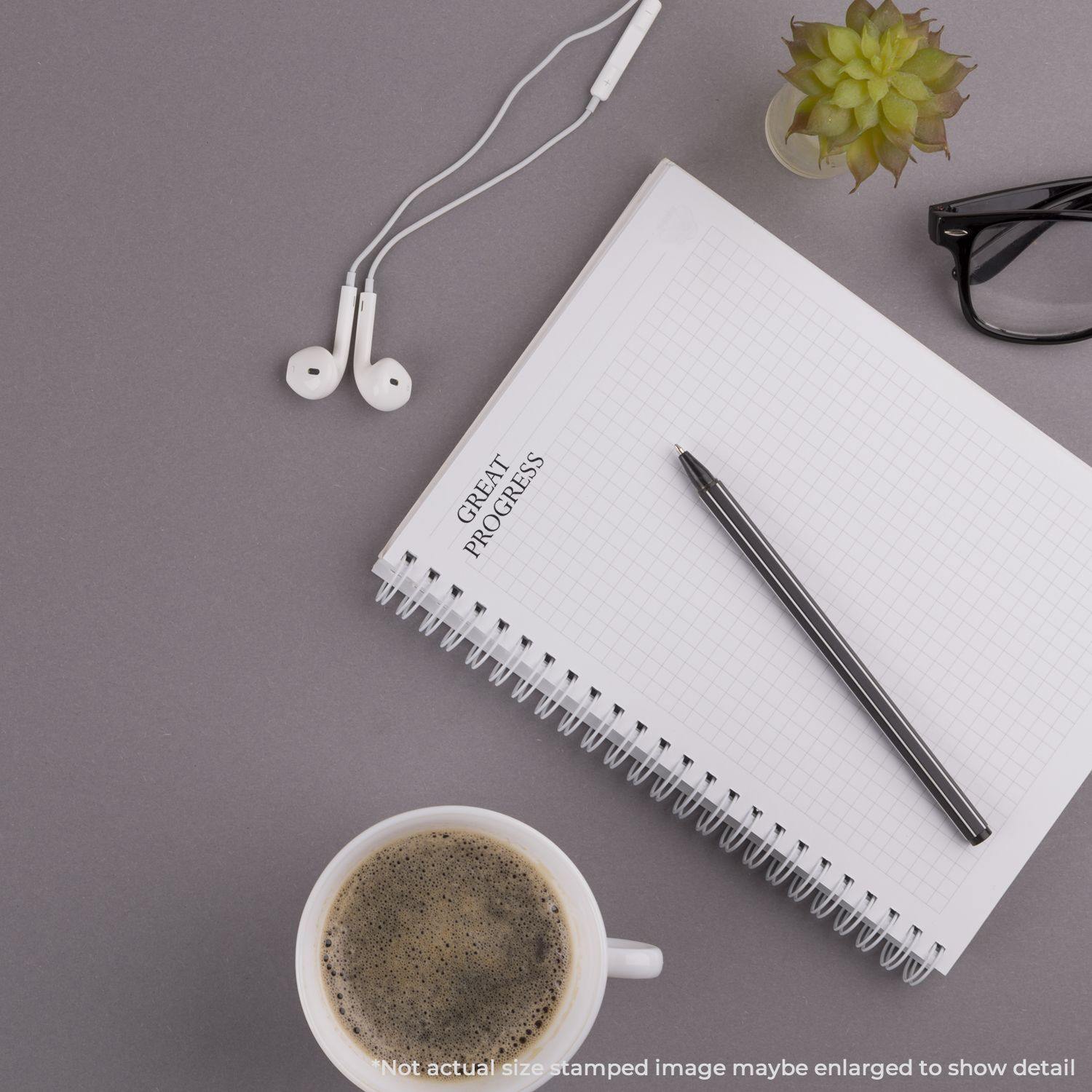 A notebook with a Self Inking Great Progress Stamp mark, pen, coffee cup, glasses, earphones, and a small plant on a gray surface.