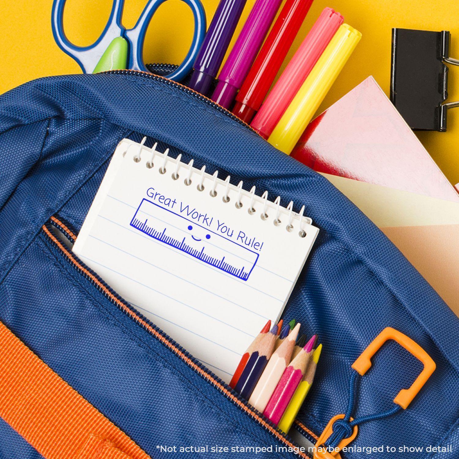 A Self Inking Great Work You Rule Stamp is used on a notepad inside a blue backpack, surrounded by colorful stationery items.