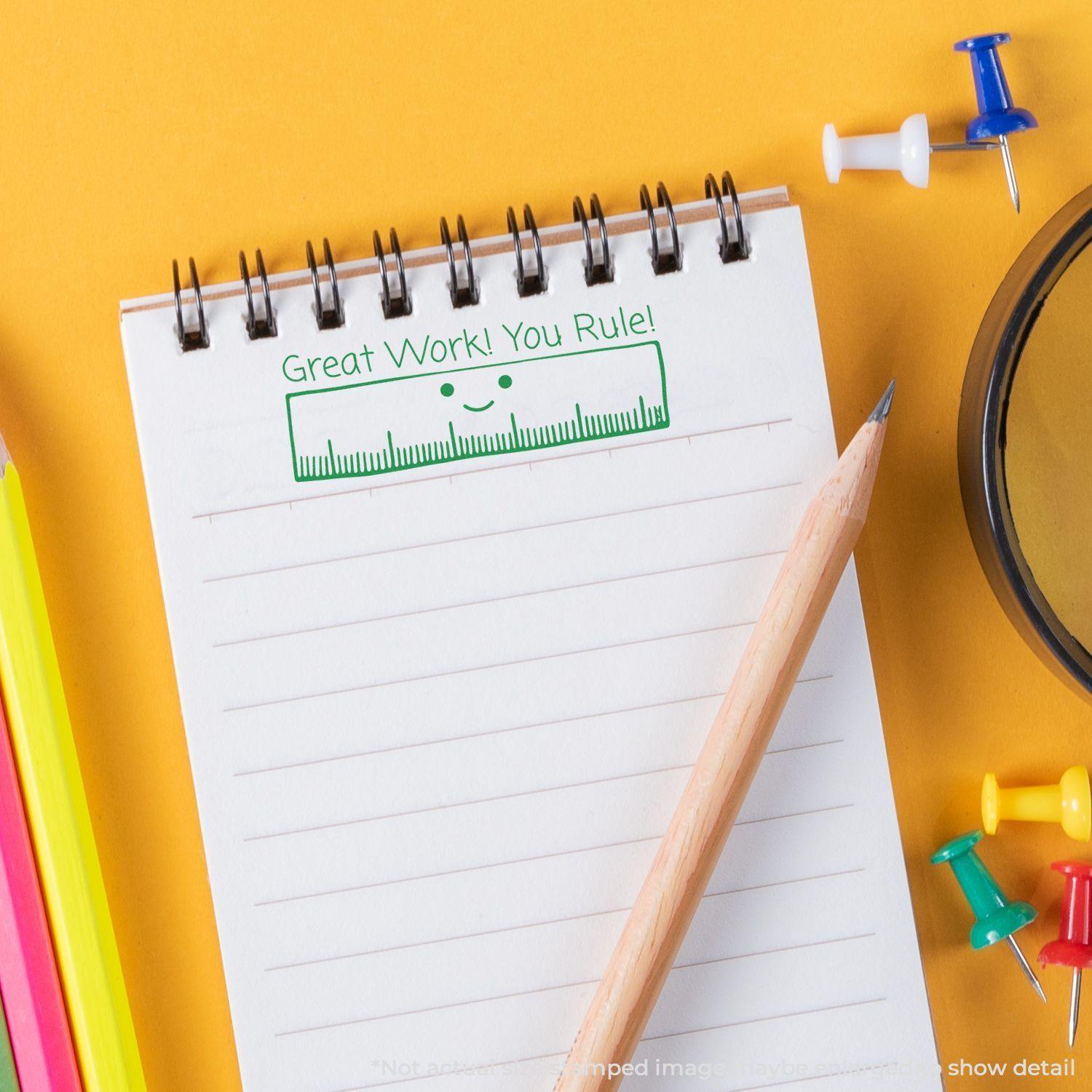 A notepad with a Great Work! You Rule rubber stamp impression, surrounded by a pencil, colorful pens, push pins, and a magnifying glass.