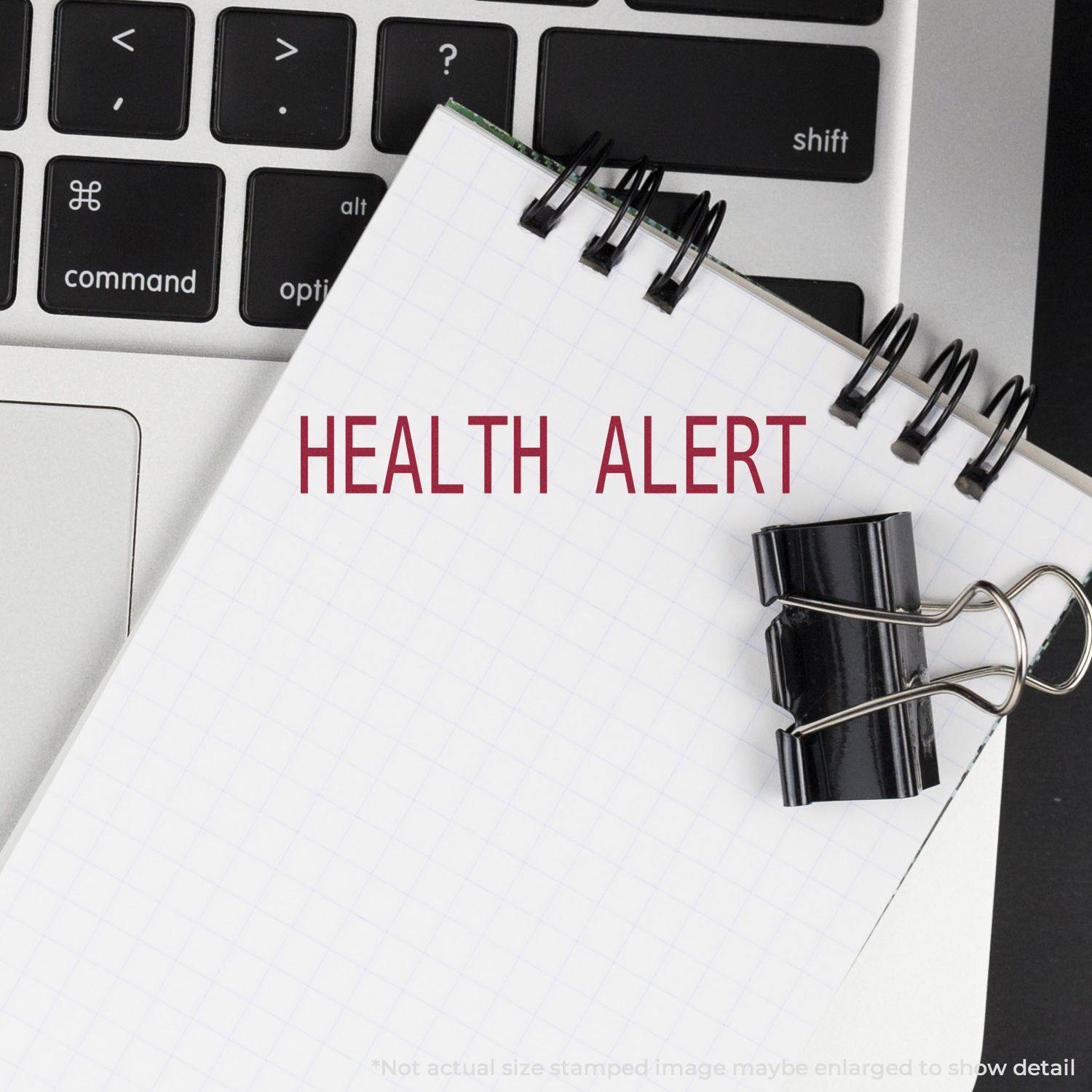 Health Alert Rubber Stamp impression on a spiral notebook placed on a laptop keyboard, with a black binder clip attached to the notebook.