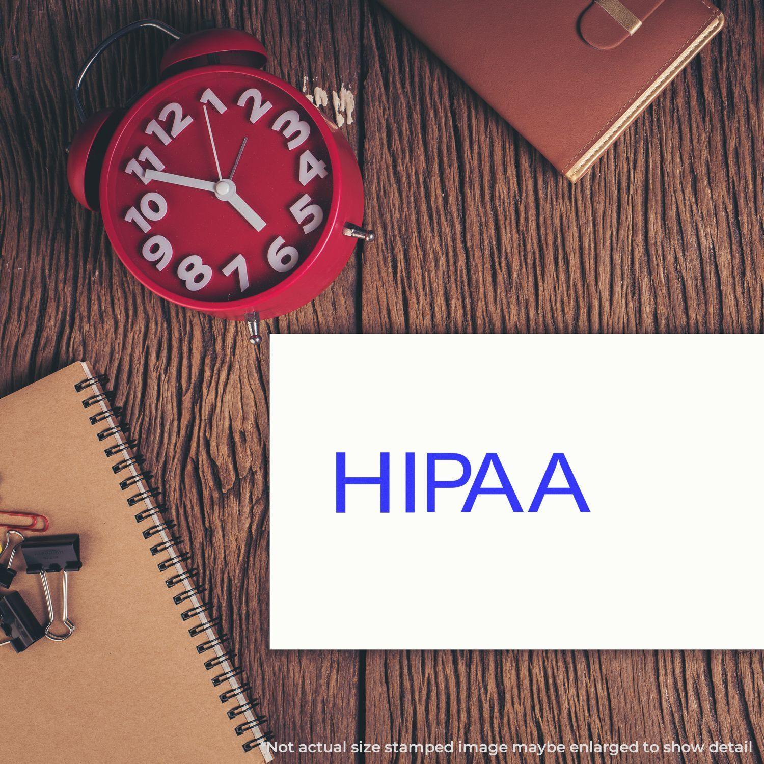 Hipaa Medical Rubber Stamp on a white card, placed on a wooden desk with a red clock, notebook, and office supplies.