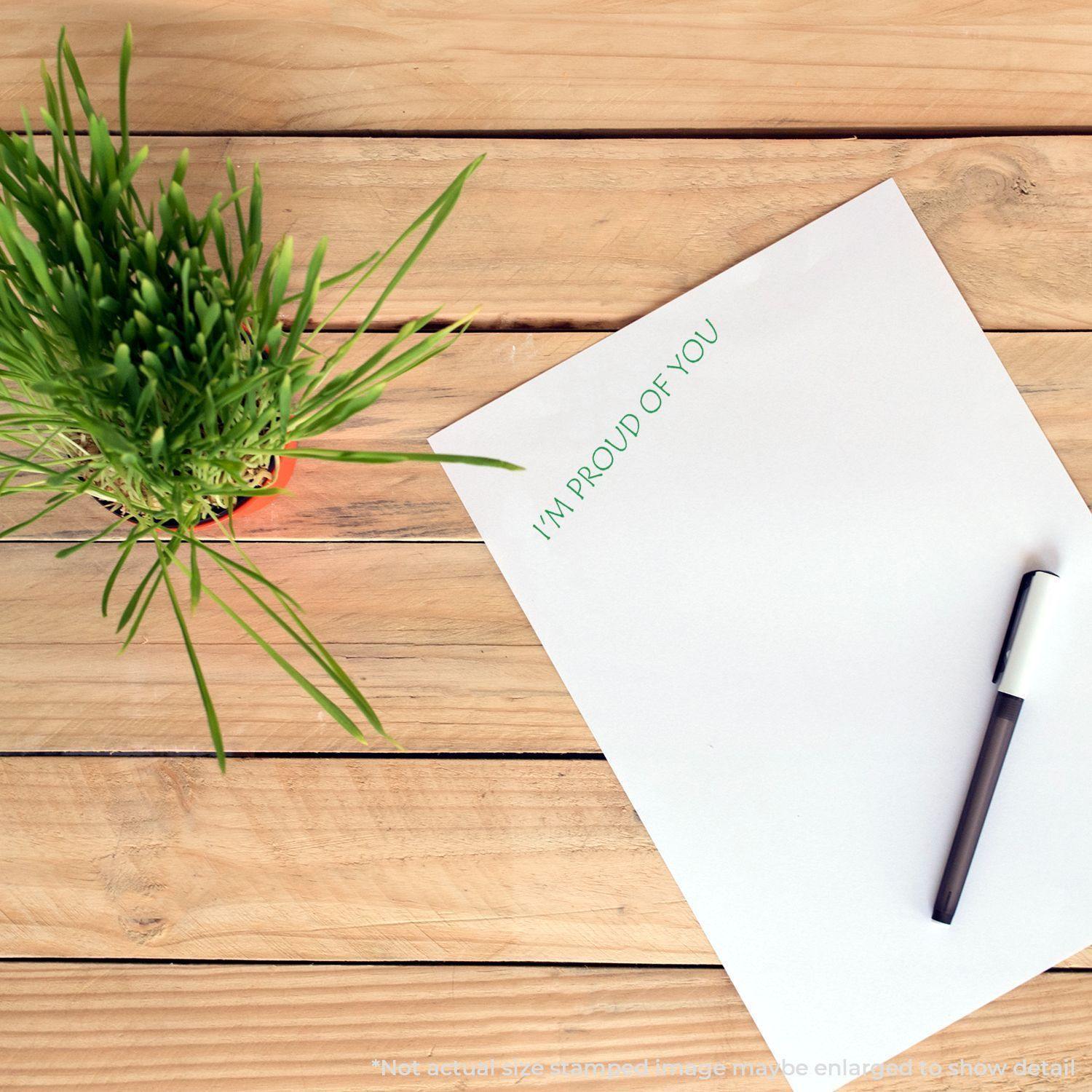 A Self Inking I'm Proud Of You Stamp is used on a white paper next to a pen and a potted plant on a wooden surface.