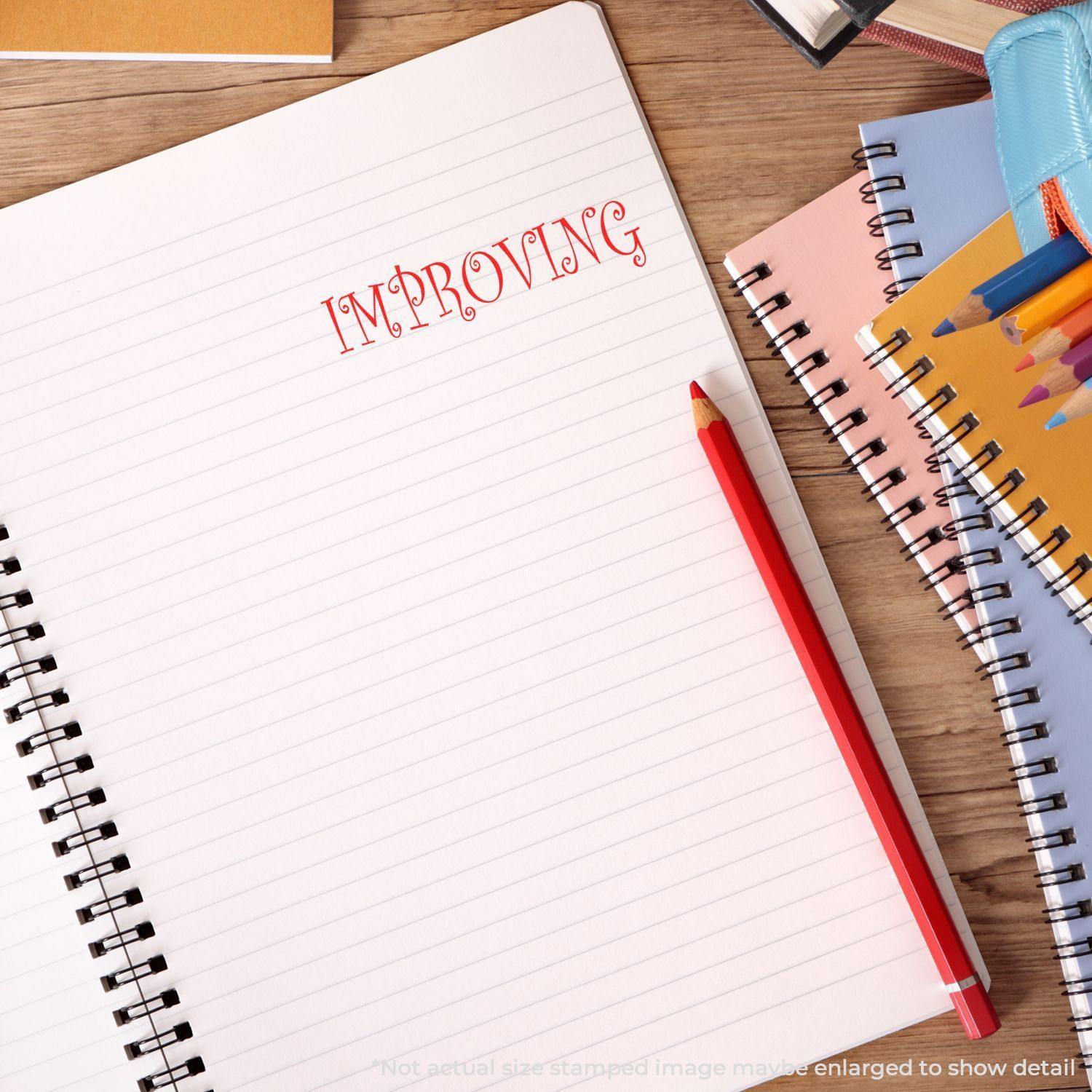 A notebook with IMPROVING stamped in red using the Large Pre-Inked Improving Stamp, surrounded by stationery items like pencils and notebooks.