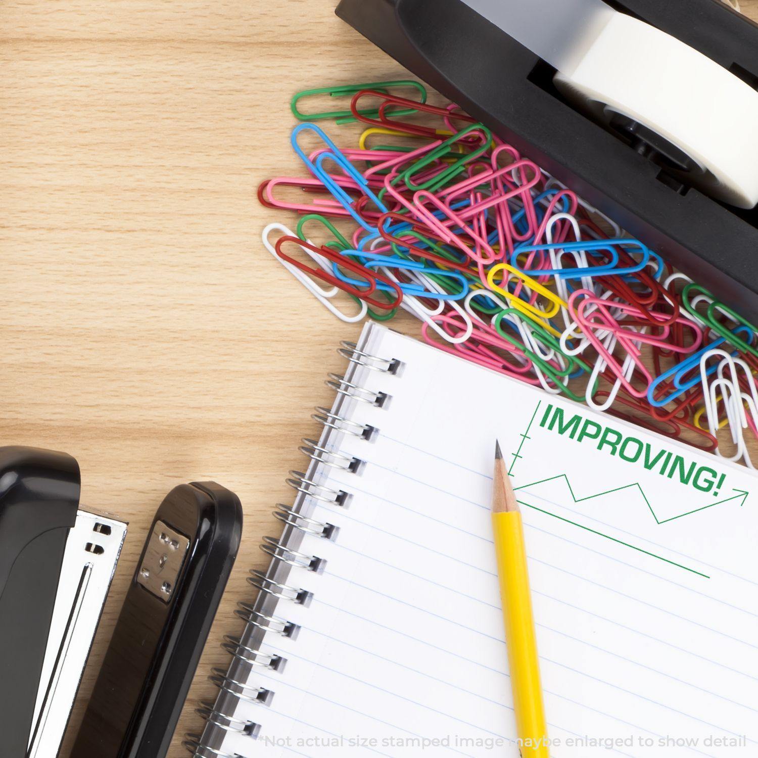 Large Self Inking Improving with Chart Icon Stamp used on a notebook page, surrounded by colorful paperclips, a stapler, and tape dispenser.