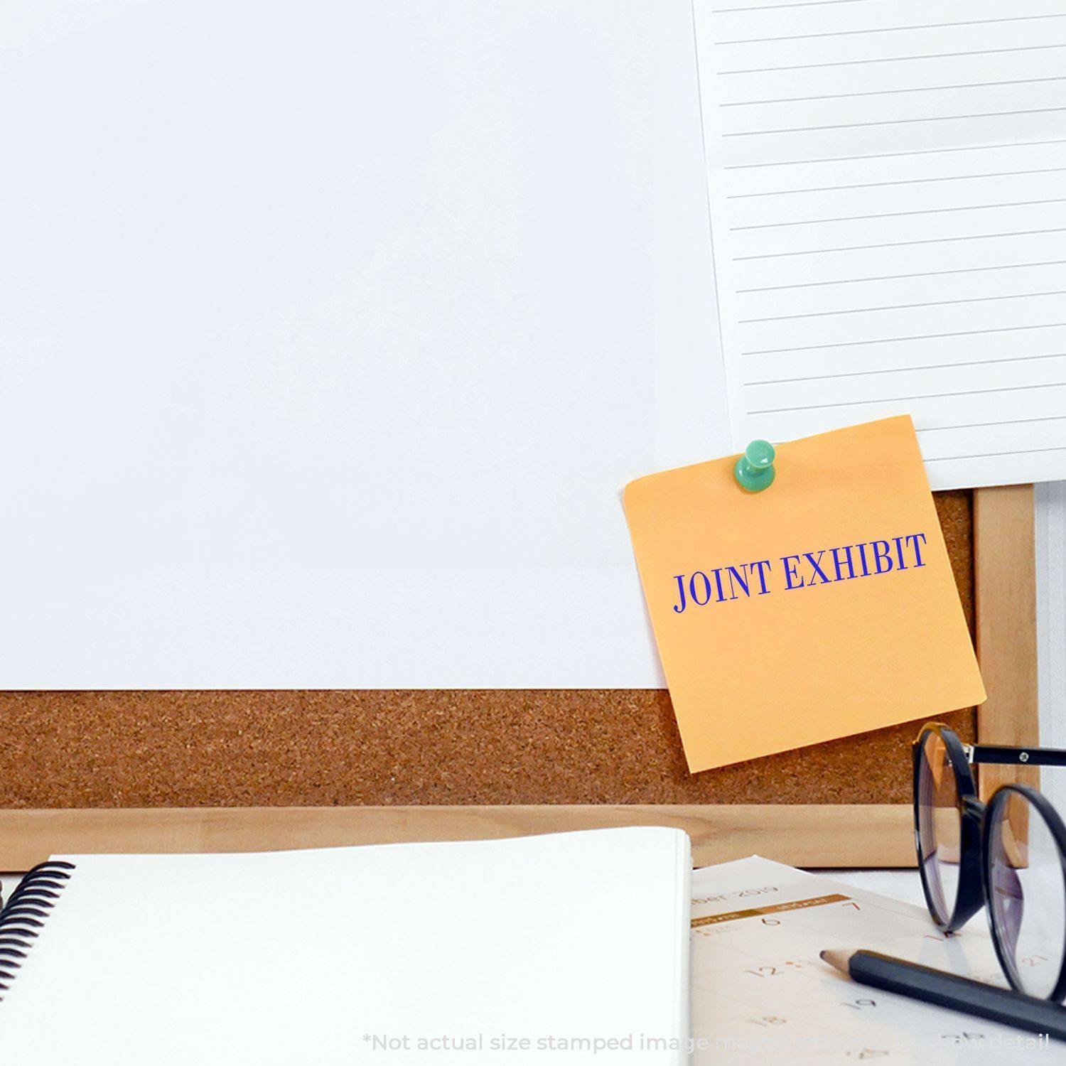 A corkboard with a note stamped JOINT EXHIBIT using the Joint Exhibit Rubber Stamp, alongside a notebook, glasses, and papers.