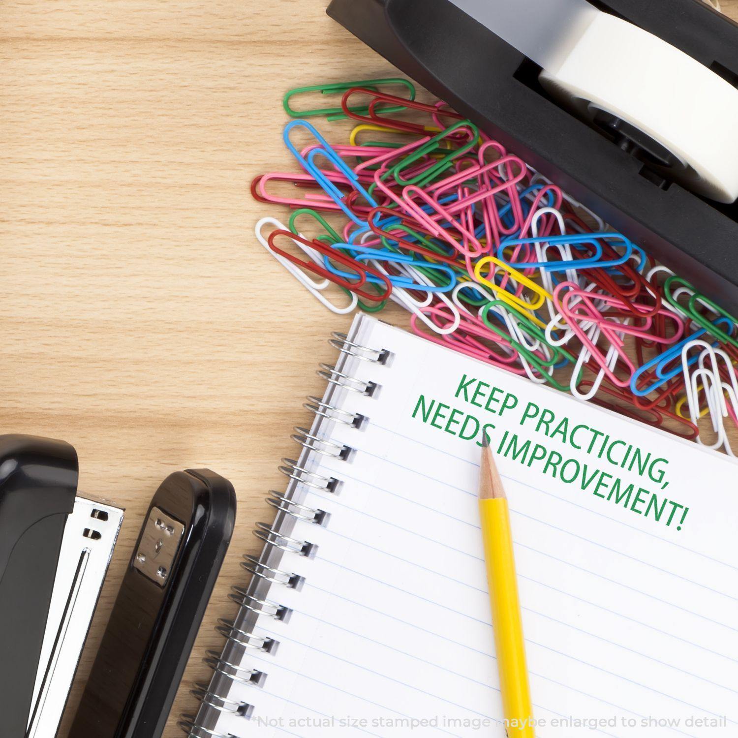 A desk with colorful paperclips, a stapler, tape dispenser, and a notebook stamped with Keep Practicing Needs Improvement Rubber Stamp in green.