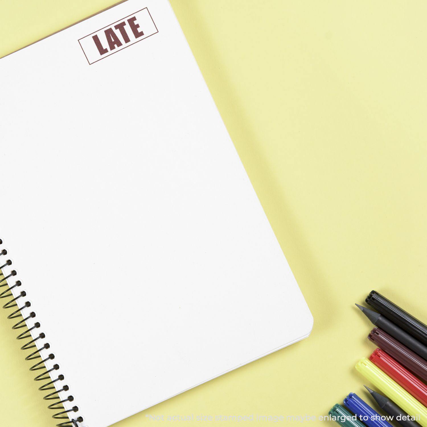 A notebook stamped with LATE using the Large Late with Border Rubber Stamp, surrounded by colorful pens on a yellow background.