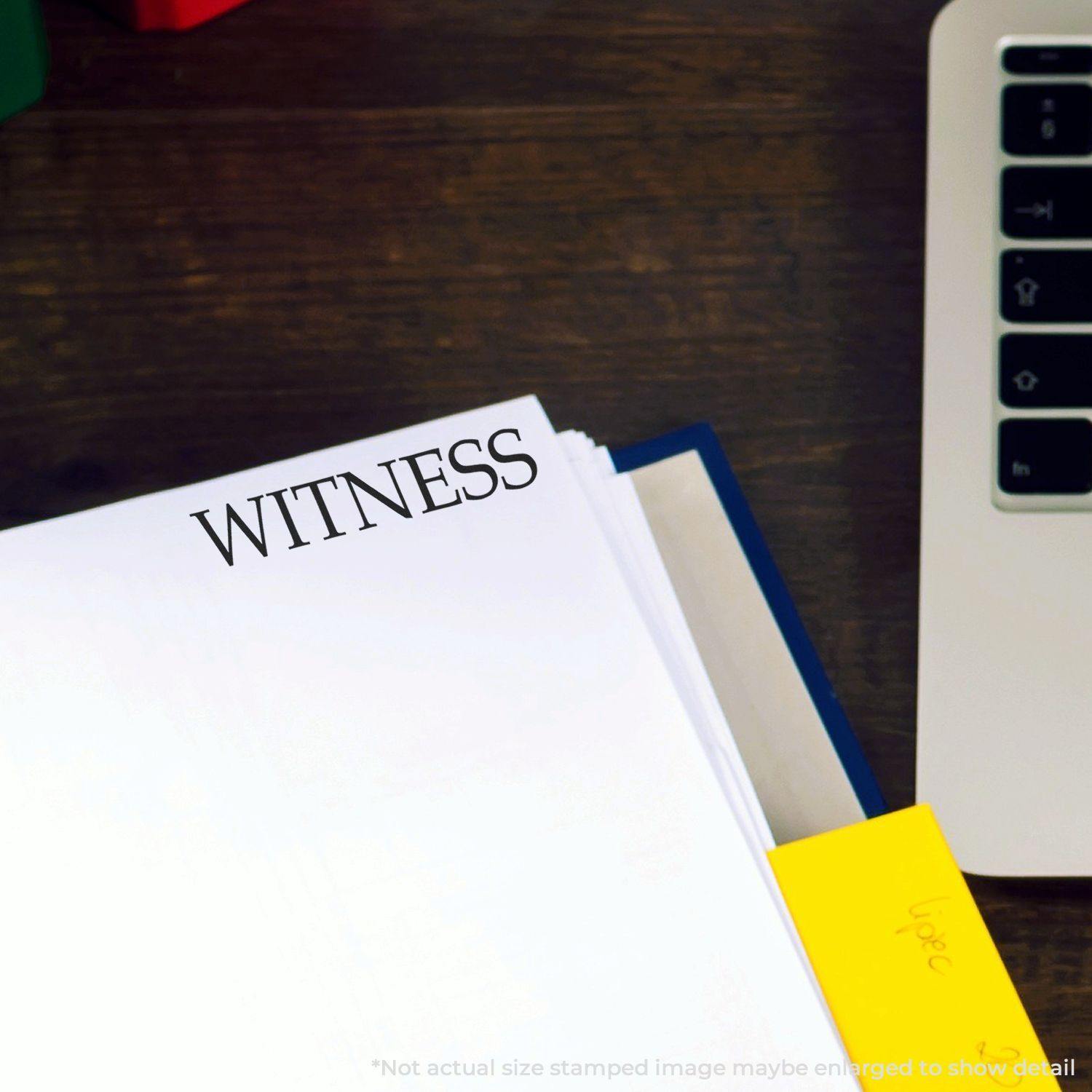 Self Inking Witness Stamp marking WITNESS on a white document near a laptop and yellow sticky note on a wooden desk.