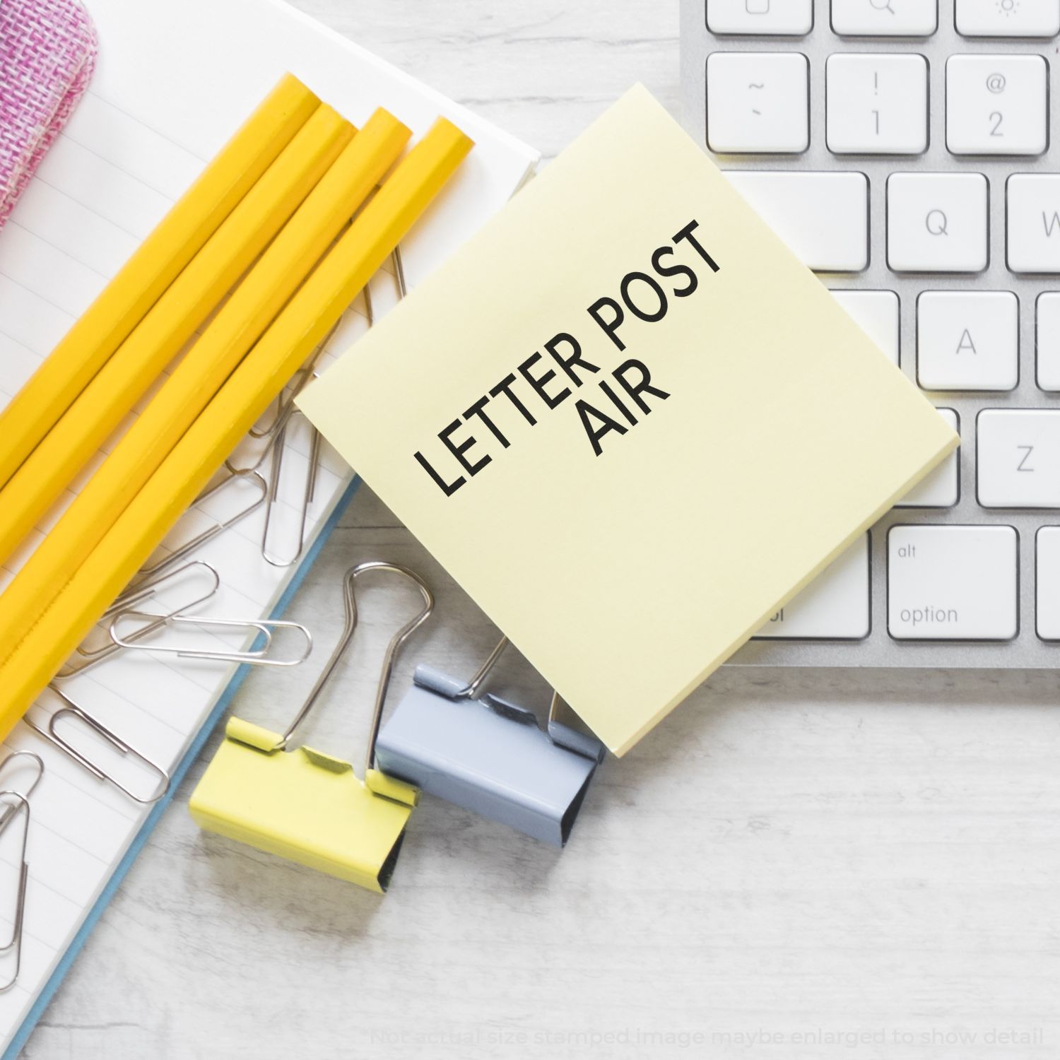 Self Inking Letter Post Air Stamp on a yellow sticky note, surrounded by pencils, paper clips, binder clips, and a keyboard.