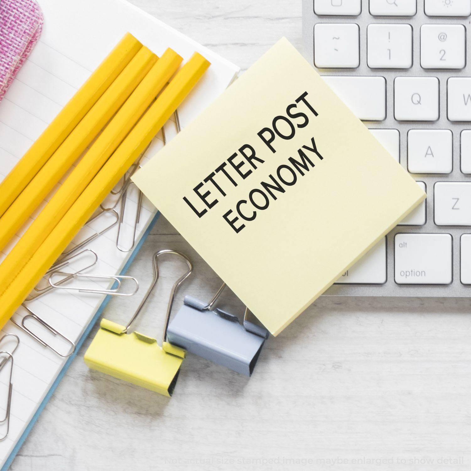 Large Letter Post Economy Rubber Stamp on a yellow sticky note, surrounded by pencils, paper clips, and a keyboard on a desk.