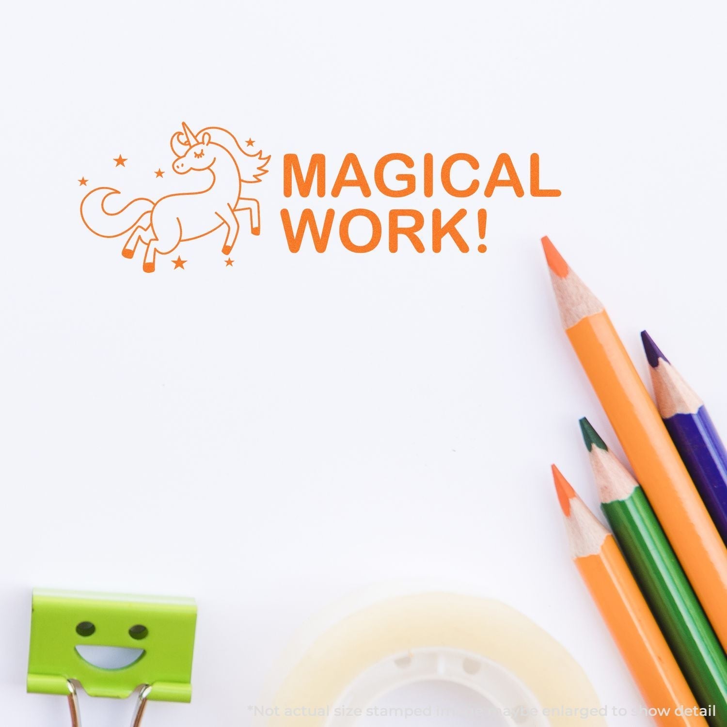 Self Inking Magical Work Stamp imprint of a unicorn and MAGICAL WORK! text in orange, shown with colored pencils, tape, and a smiling binder clip.