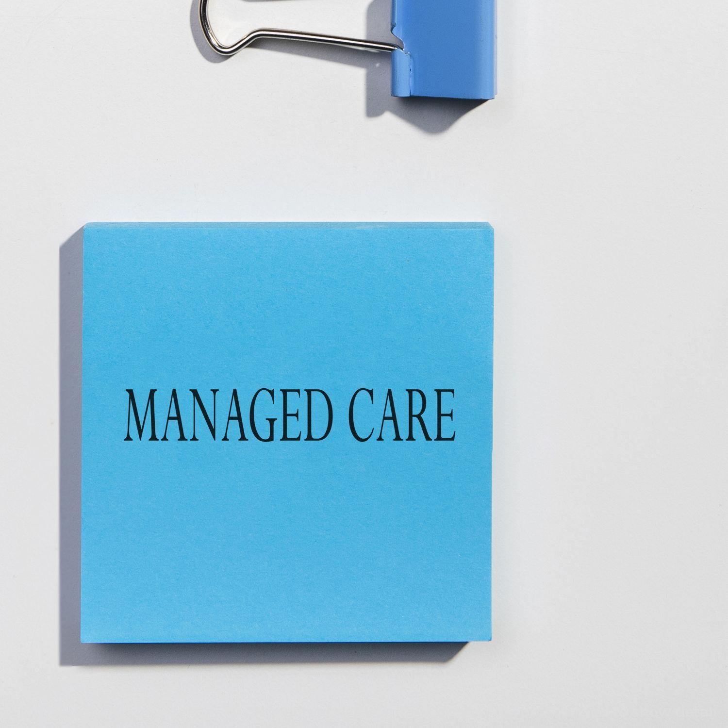 Blue Slim Pre-Inked Managed Care Stamp on a white surface with a blue binder clip in the background.