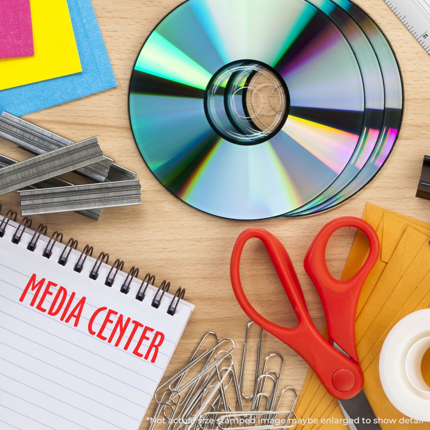 Self Inking Media Center Stamp on a notepad, surrounded by CDs, red scissors, paper clips, staples, and colorful paper on a wooden desk.