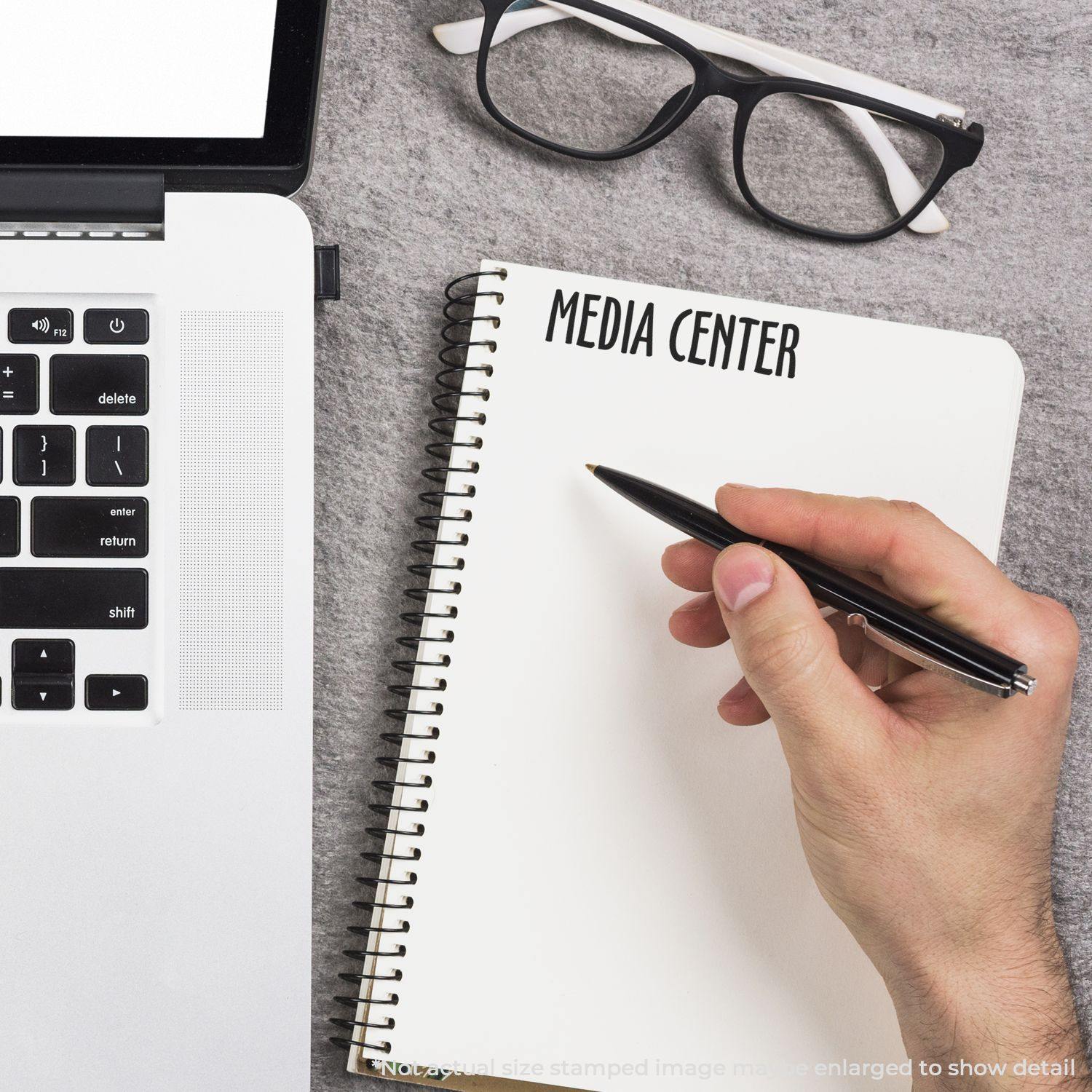 A hand writing in a notebook stamped with MEDIA CENTER using the Large Media Center Rubber Stamp, next to a laptop and glasses.