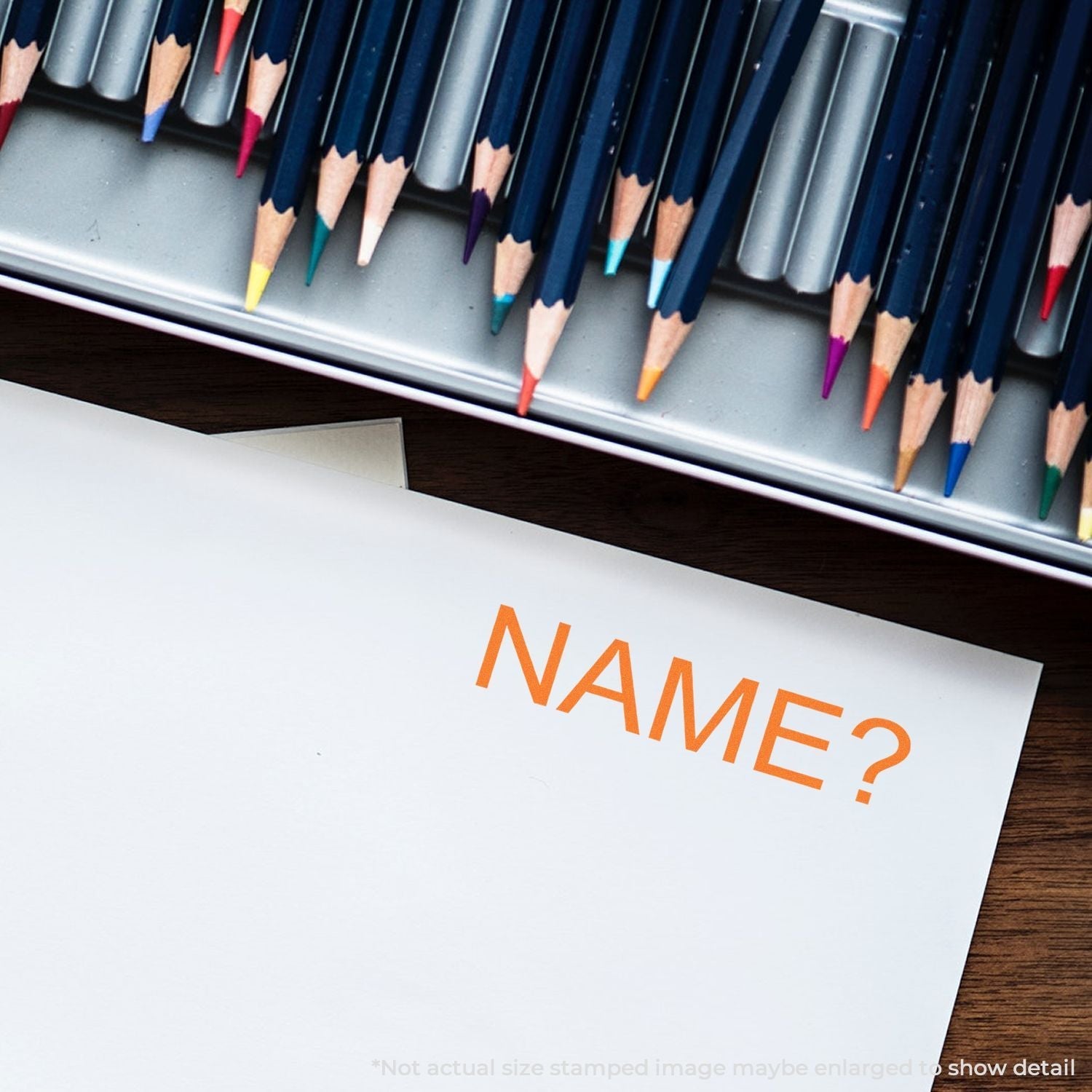 Self Inking Name Stamp in use on white paper, with colored pencils in the background. The stamp prints NAME? in orange ink.