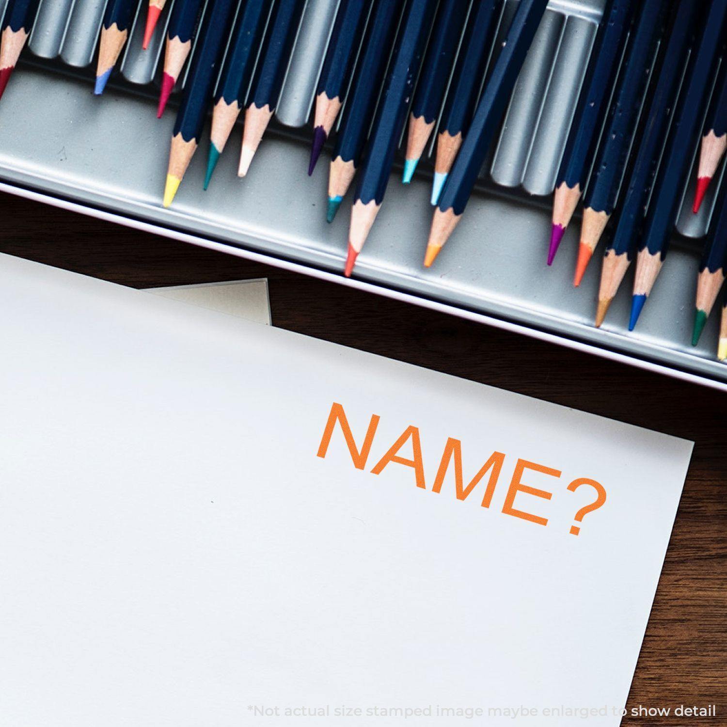 Name Rubber Stamp in use on white paper, with a tray of colored pencils in the background.