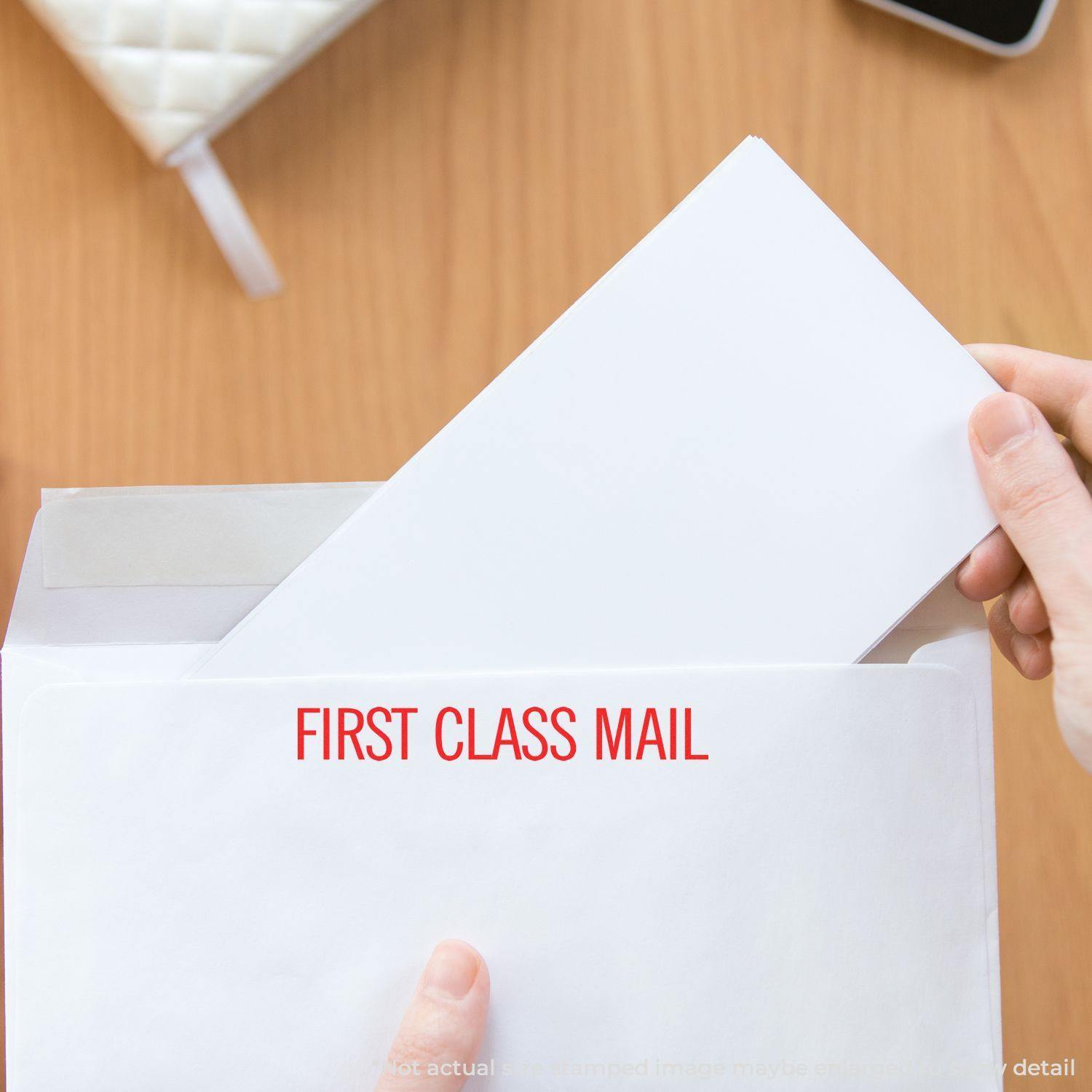 Person holding an envelope stamped with FIRST CLASS MAIL using the Large Narrow First Class Mail Rubber Stamp on a wooden table.