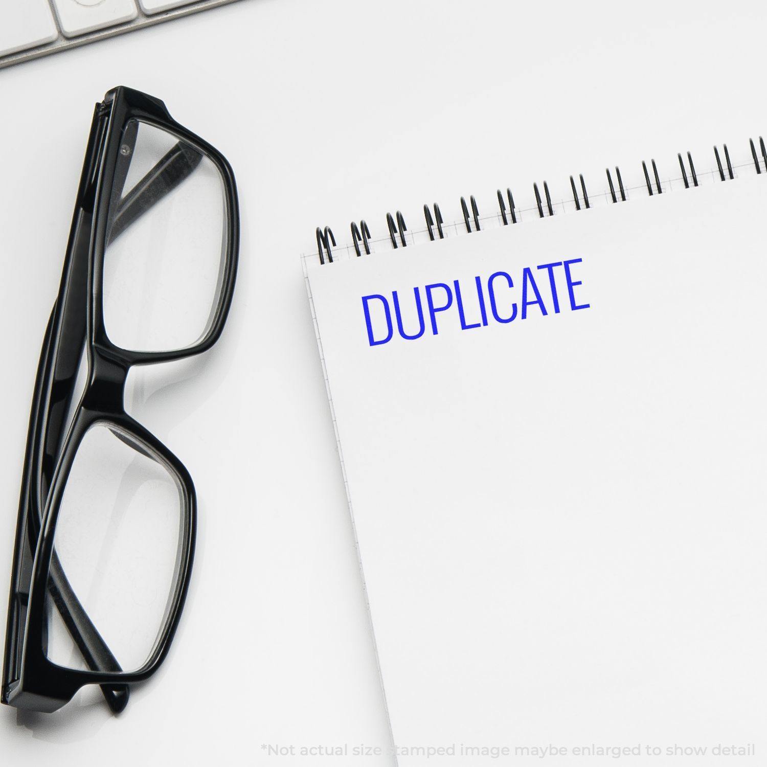 Narrow Font Duplicate Rubber Stamp used on a white notepad, with black glasses and a keyboard in the background.