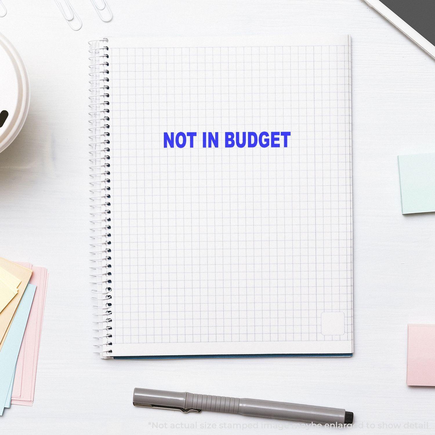 A notebook with a Not In Budget rubber stamp imprint in blue ink on a grid page, surrounded by office supplies.