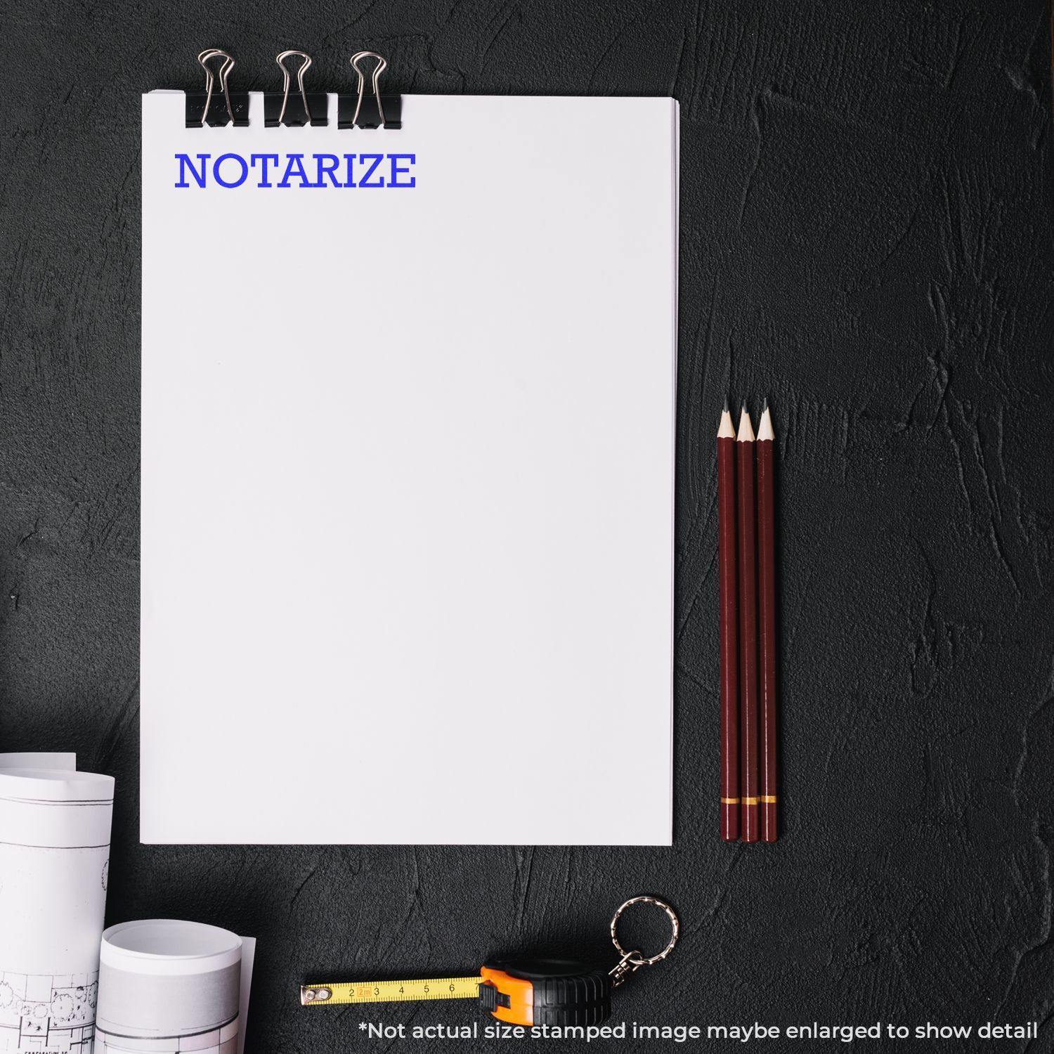 Large Notarize Rubber Stamp in blue ink on a white paper with binder clips, pencils, measuring tape, and rolled blueprints on a black surface.