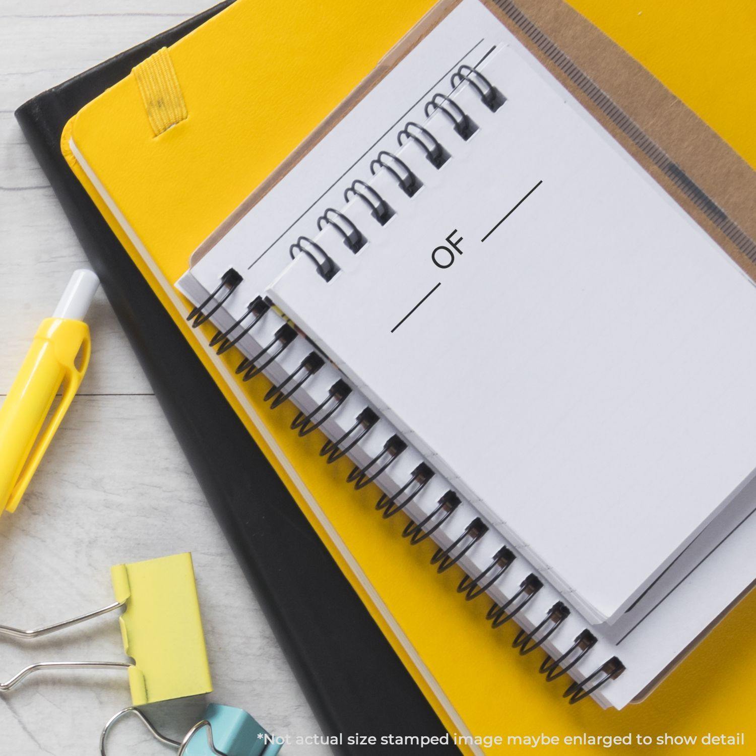 A Large Of Rubber Stamp is shown in use on a spiral notebook, surrounded by yellow and black notebooks, a pen, and binder clips.