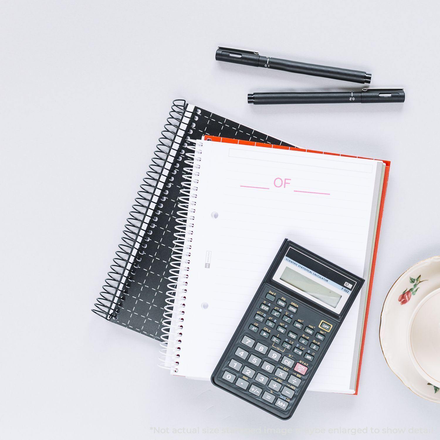 A rubber stamp with the word OF is stamped on a white notebook page, surrounded by pens, a calculator, and a teacup.