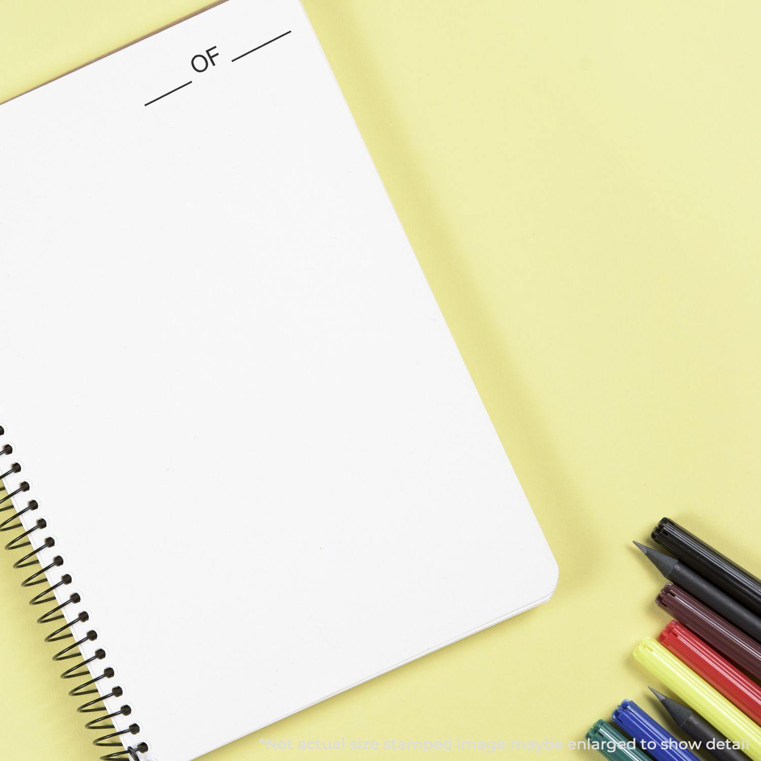 A Large Of Rubber Stamp is used on a spiral notebook with OF stamped at the top. Pens and highlighters are placed beside the notebook.