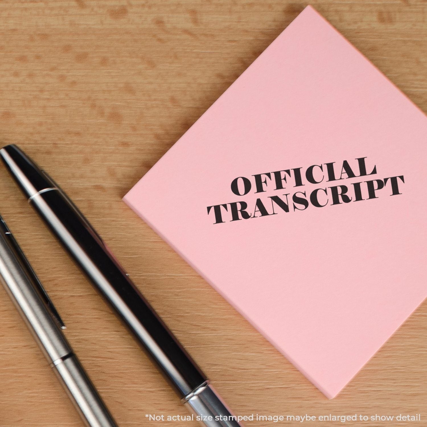 Self Inking Official Transcript Stamp on a pink paper beside two pens on a wooden surface. Text at bottom notes image is enlarged to show detail.