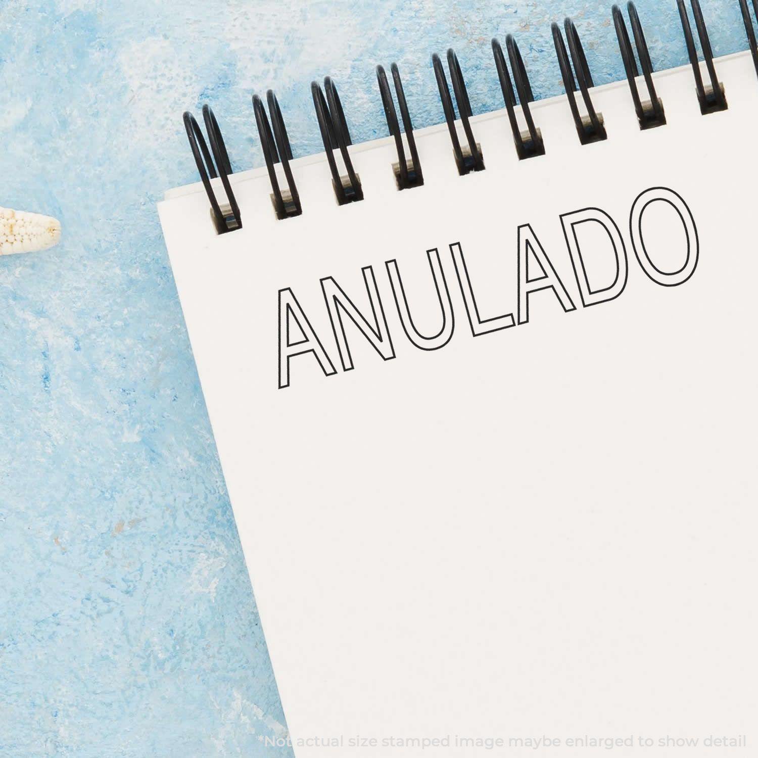 A notebook with the word ANULADO stamped on it using the Large Outline Anulado Rubber Stamp, placed on a blue textured surface.