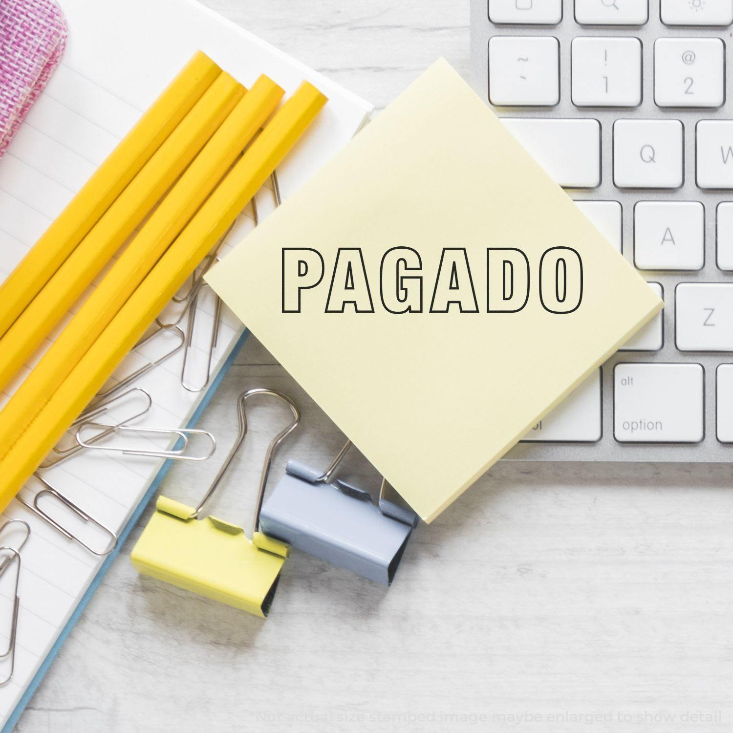 Large Outline Pagado Rubber Stamp on a yellow sticky note, placed on a desk with pencils, paper clips, binder clips, and a keyboard.