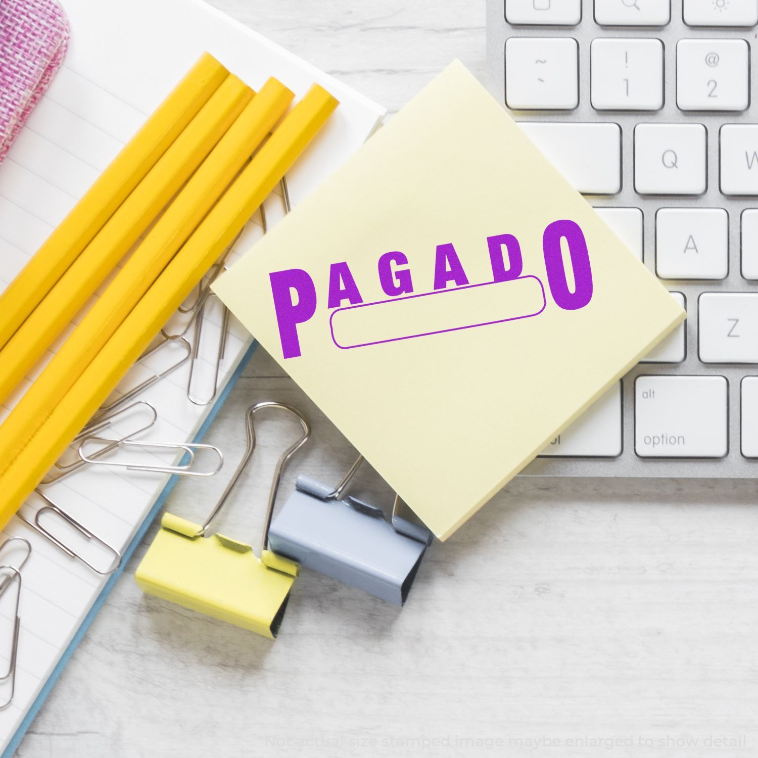 Self Inking Pagado with Box Stamp in use on a sticky note, placed on a desk with a keyboard, pencils, and paper clips.