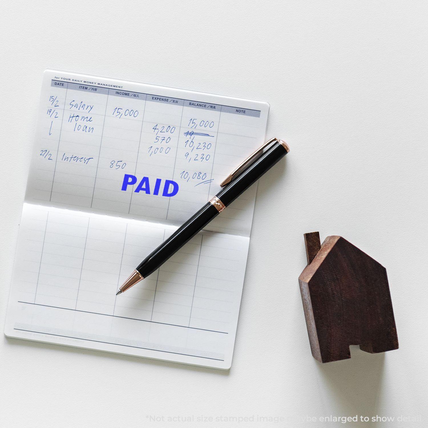 Slim Pre-Inked Paid Stamp on a checkbook with a pen and wooden house-shaped object on a white surface.