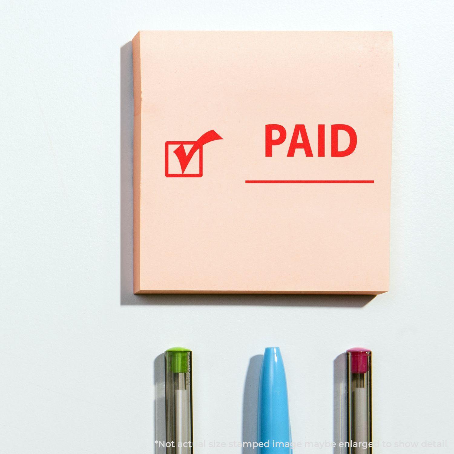 Large Paid with Checkmark Rubber Stamp used on a pink sticky note with three pens below it.