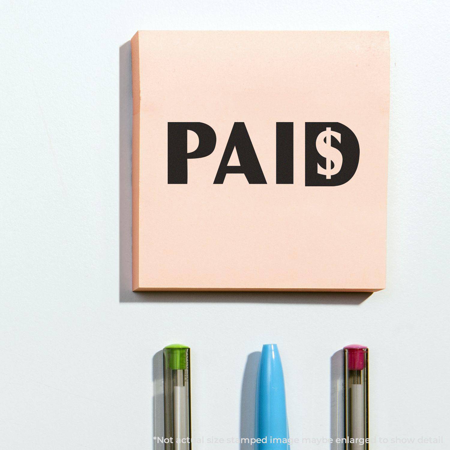 Large Paid with Dollar Sign Rubber Stamp used on a pink sticky note, with three pens below it on a white surface.