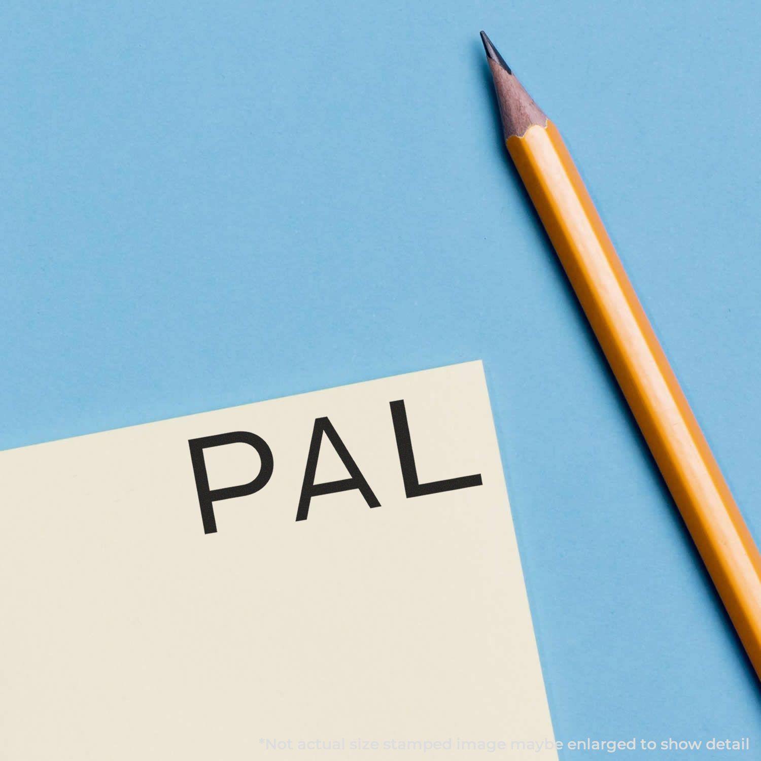 A Large PAL Rubber Stamp imprint on a yellow paper next to a sharpened yellow pencil on a blue background.