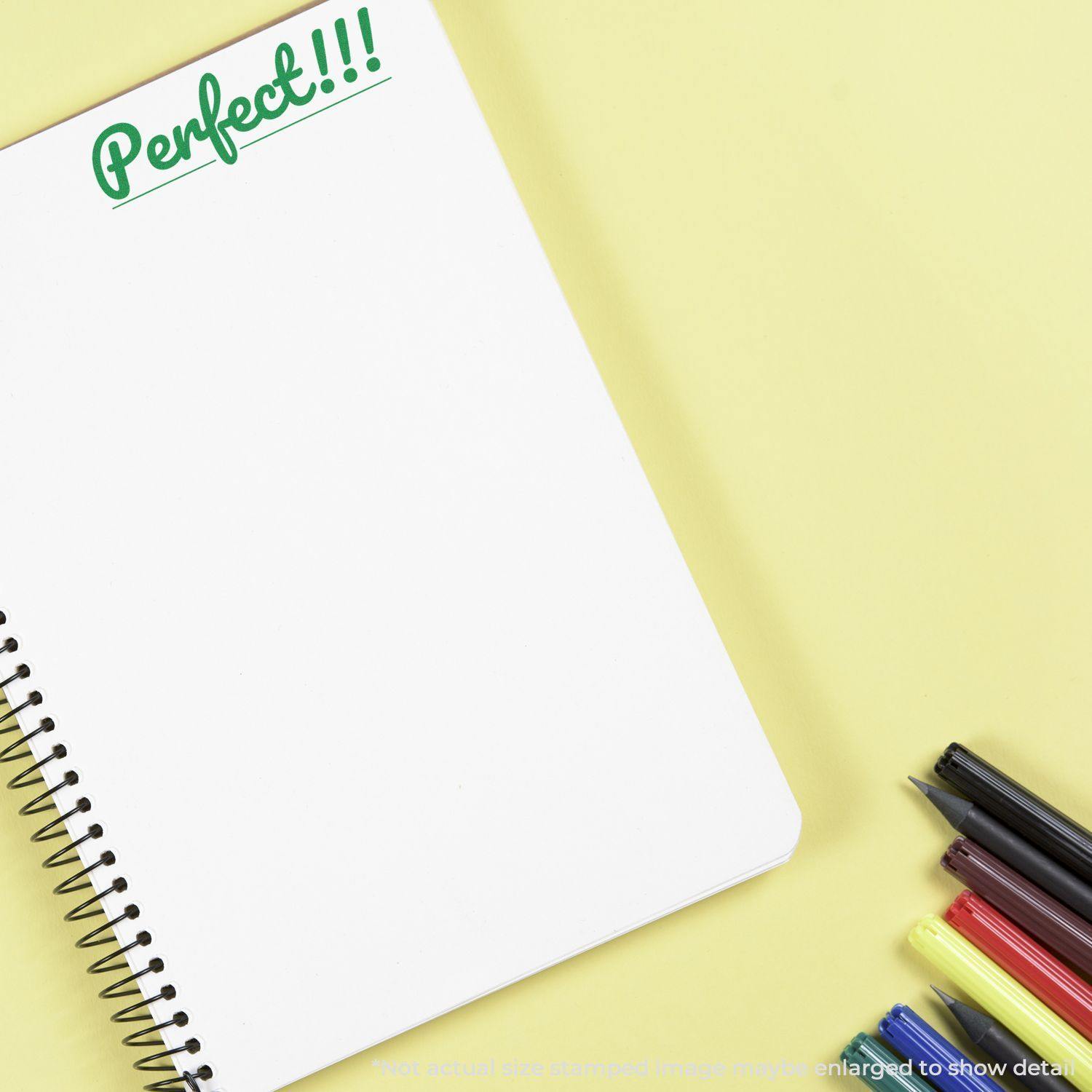 A Large Perfect Rubber Stamp imprinting Perfect!!! in green ink on a blank spiral notebook, with colorful pens beside it on a yellow background.