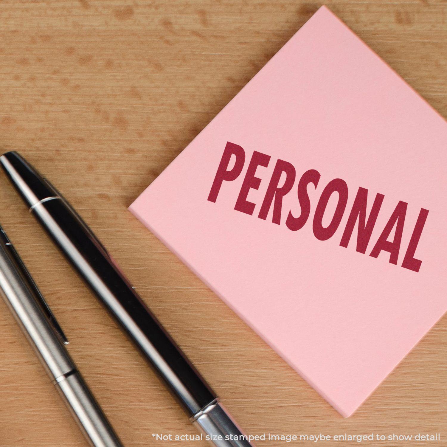 Large Self Inking Personal Stamp used on a pink sticky note with the word PERSONAL stamped in red, next to two pens on a wooden surface.