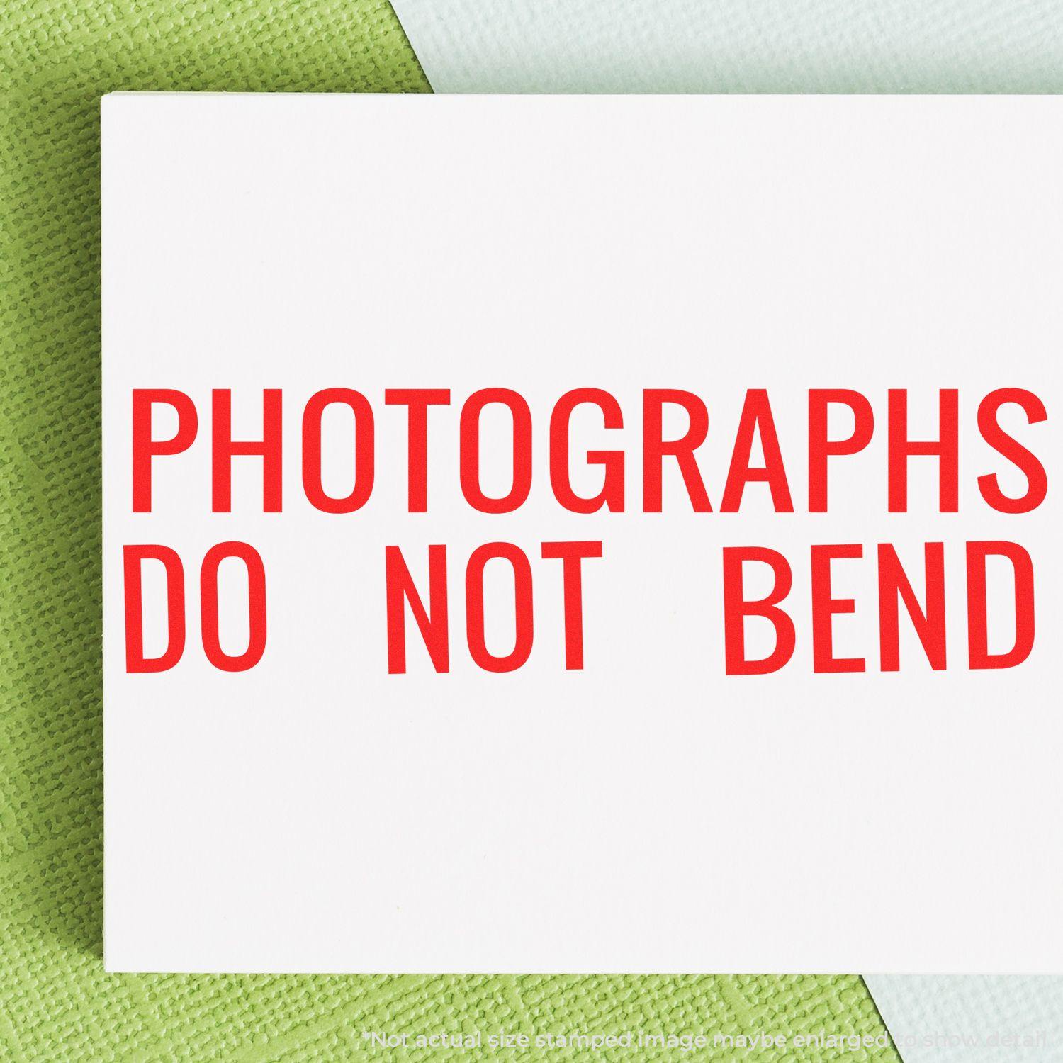 Large Photographs Do Not Bend Rubber Stamp in red ink on a white envelope, placed on a green and white textured background.