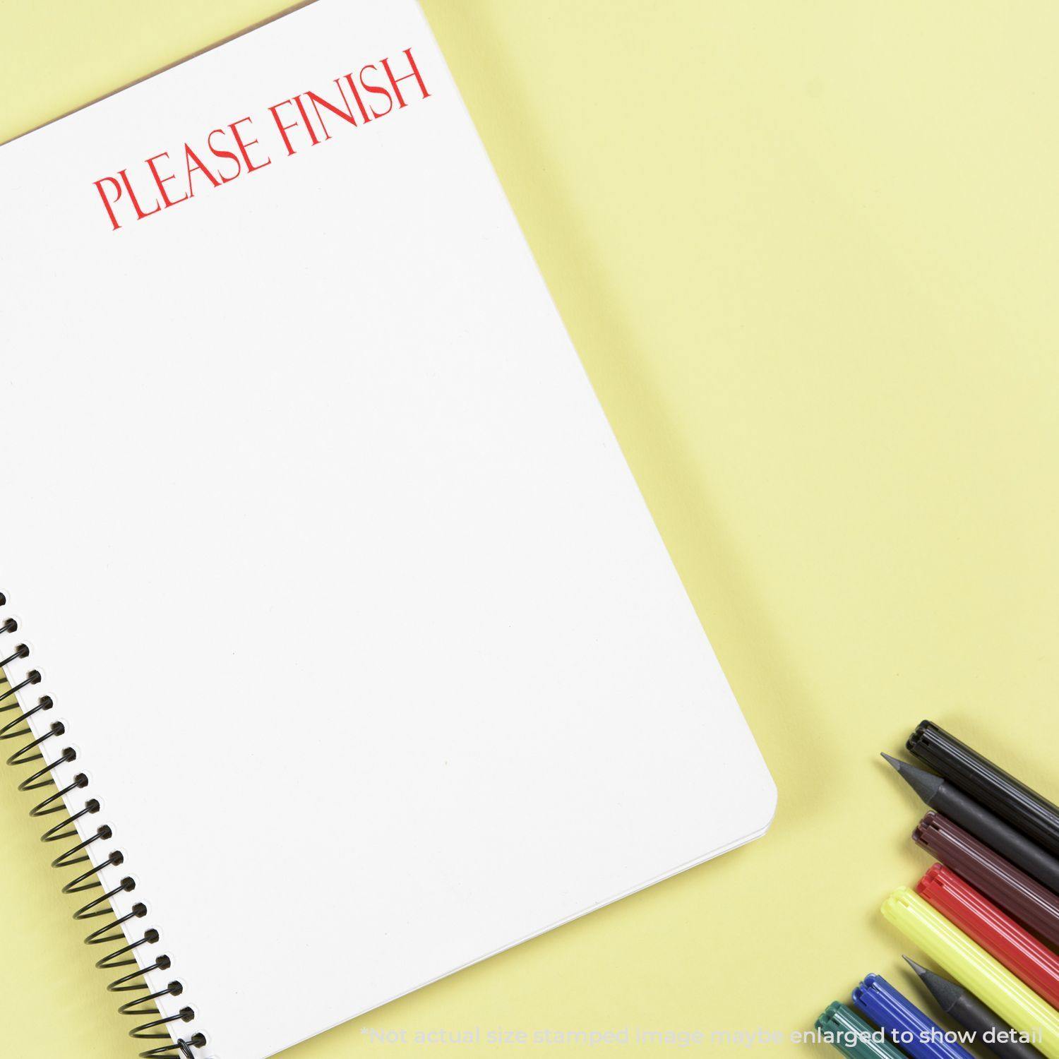 A notebook with Please Finish rubber stamp in red ink on a yellow background, next to colored pens and markers.